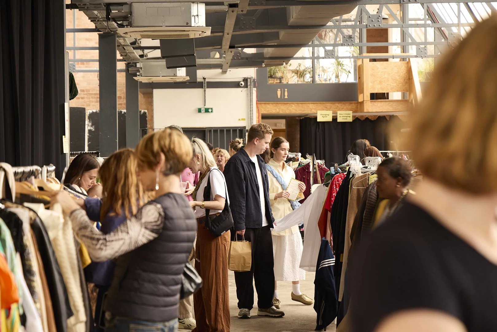 People shopping at a clothing store with racks of clothes, some browsing and others inspecting items, inside a well-lit space with high ceilings and industrial design elements.
