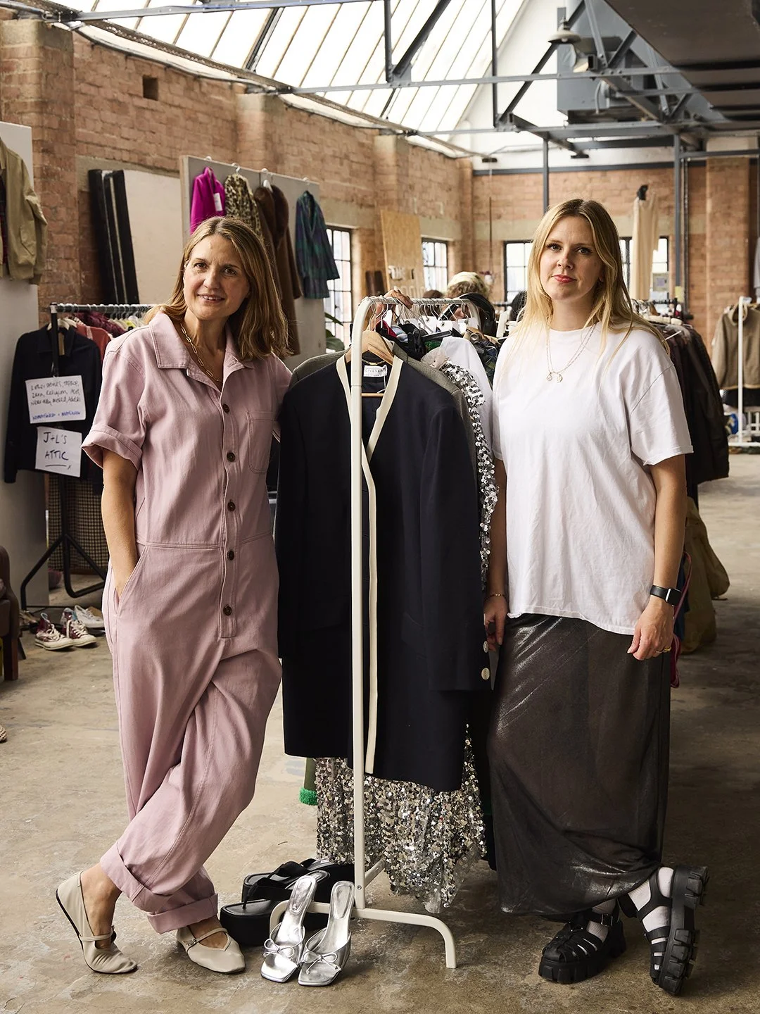Two women standing in a clothing store with racks of clothes behind them, one wearing a pink jumpsuit and the other in a white t-shirt and long skirt, surrounded by fashion items.