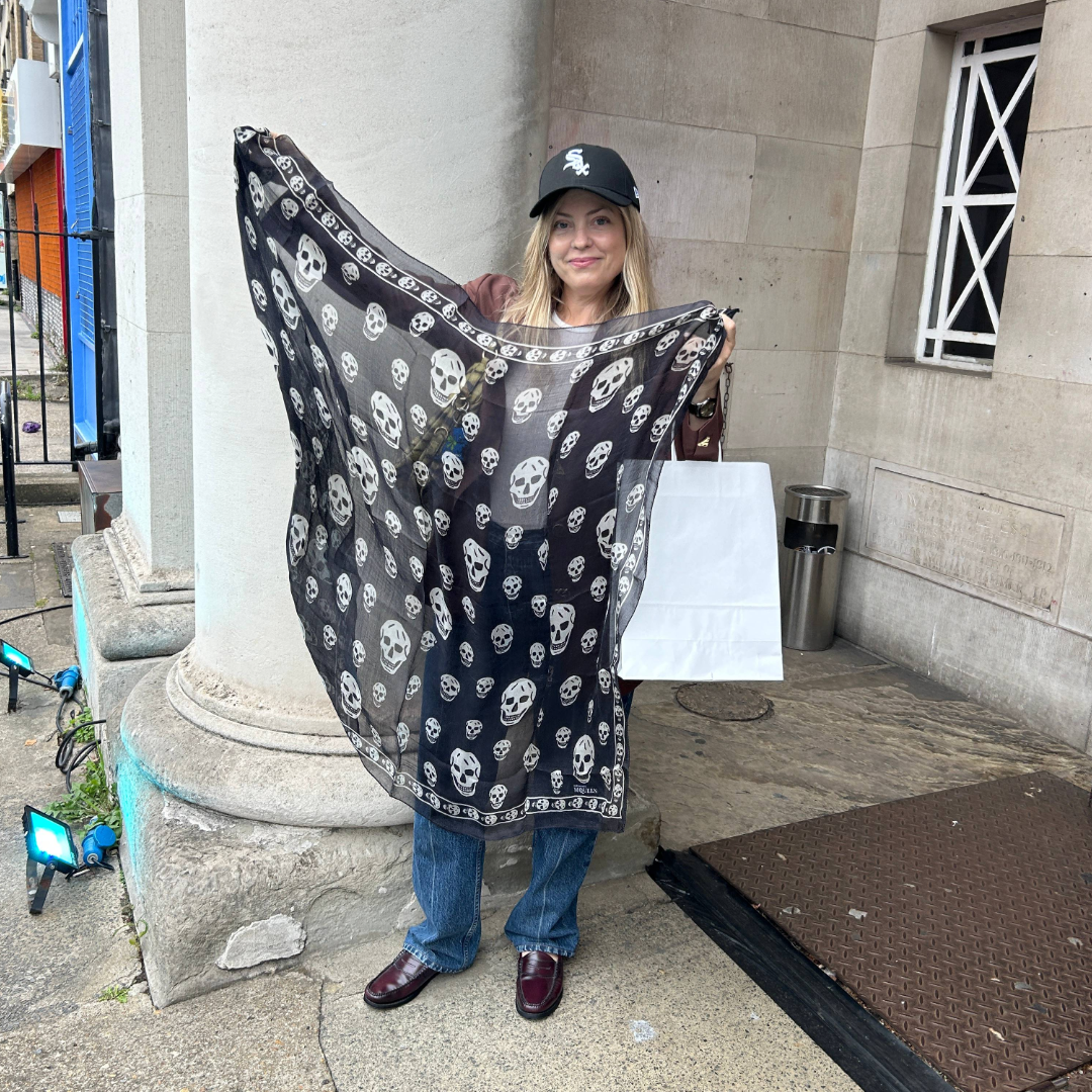 A woman holding up a sheer black scarf with white skull patterns while standing outside near a stone pillar, wearing a black cap, brown jacket, blue jeans, and burgundy shoes.