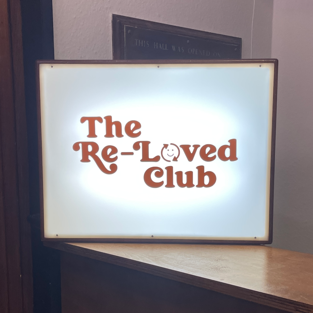 Backlit sign displaying 'The Re-Loved Club' in brown text with a smiling face in the 'O' of 'Loved'.