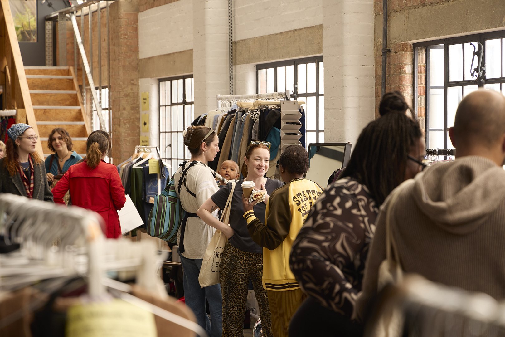 People shopping at a thrift store with racks of clothes and large windows in the background.