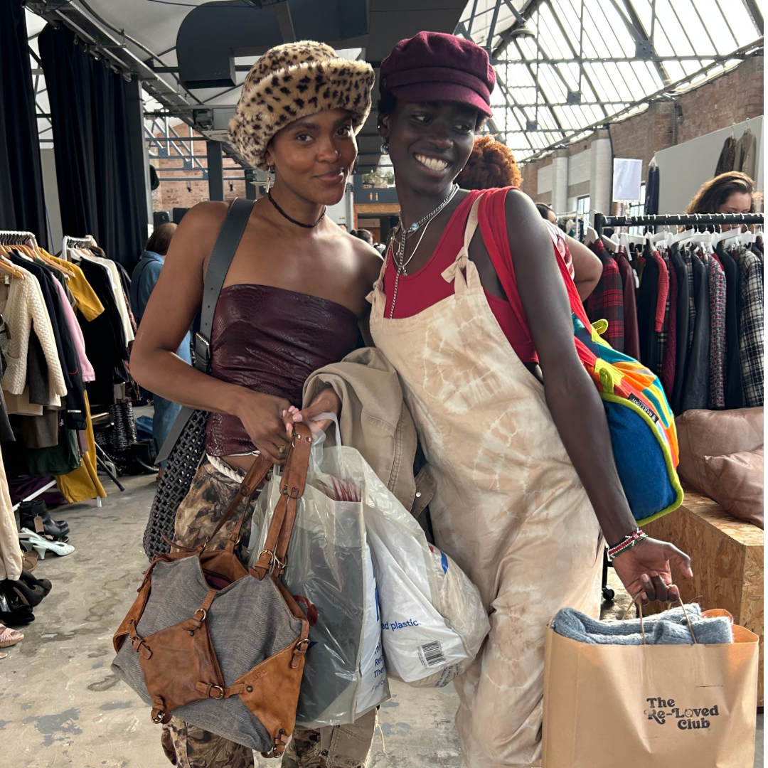 Two women shopping at a clothing market, carrying multiple shopping bags and wearing casual stylish outfits, smiling in a well-lit space with clothing racks in the background.