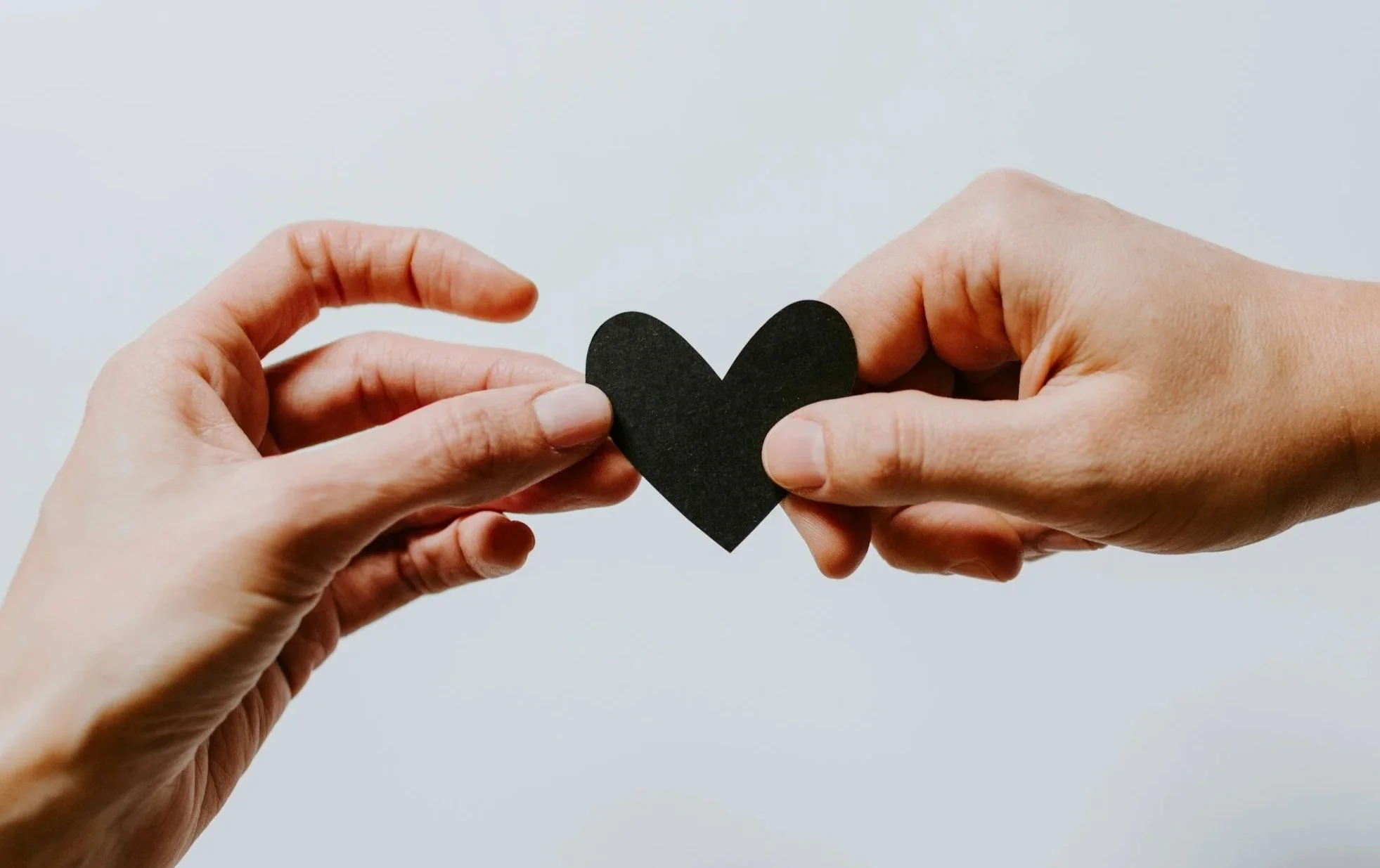 Two hands exchanging a black paper heart against a white background.