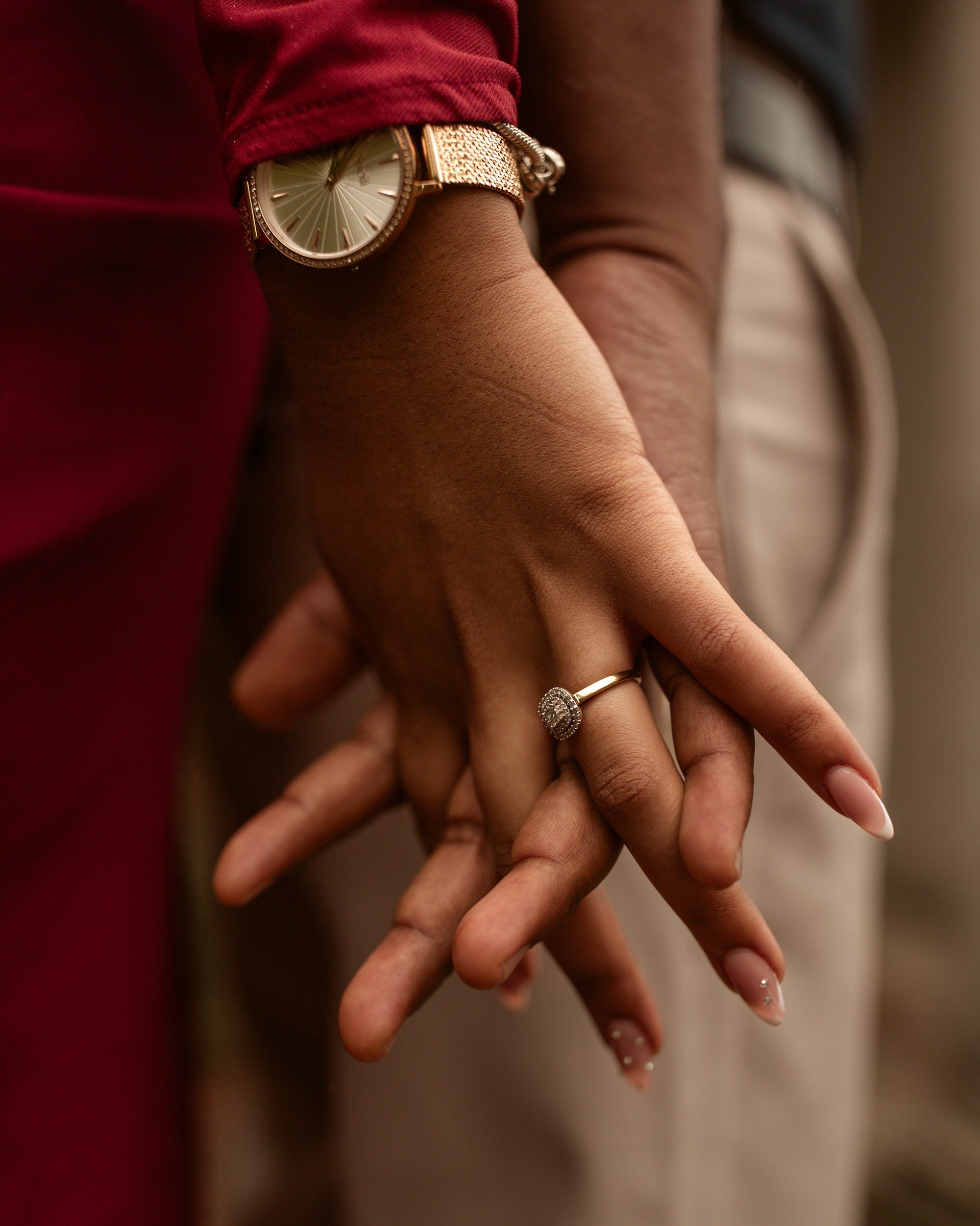 Close-up of two hands intertwined, one with a gold watch and a diamond ring, the other with manicured nails with small embellishments.