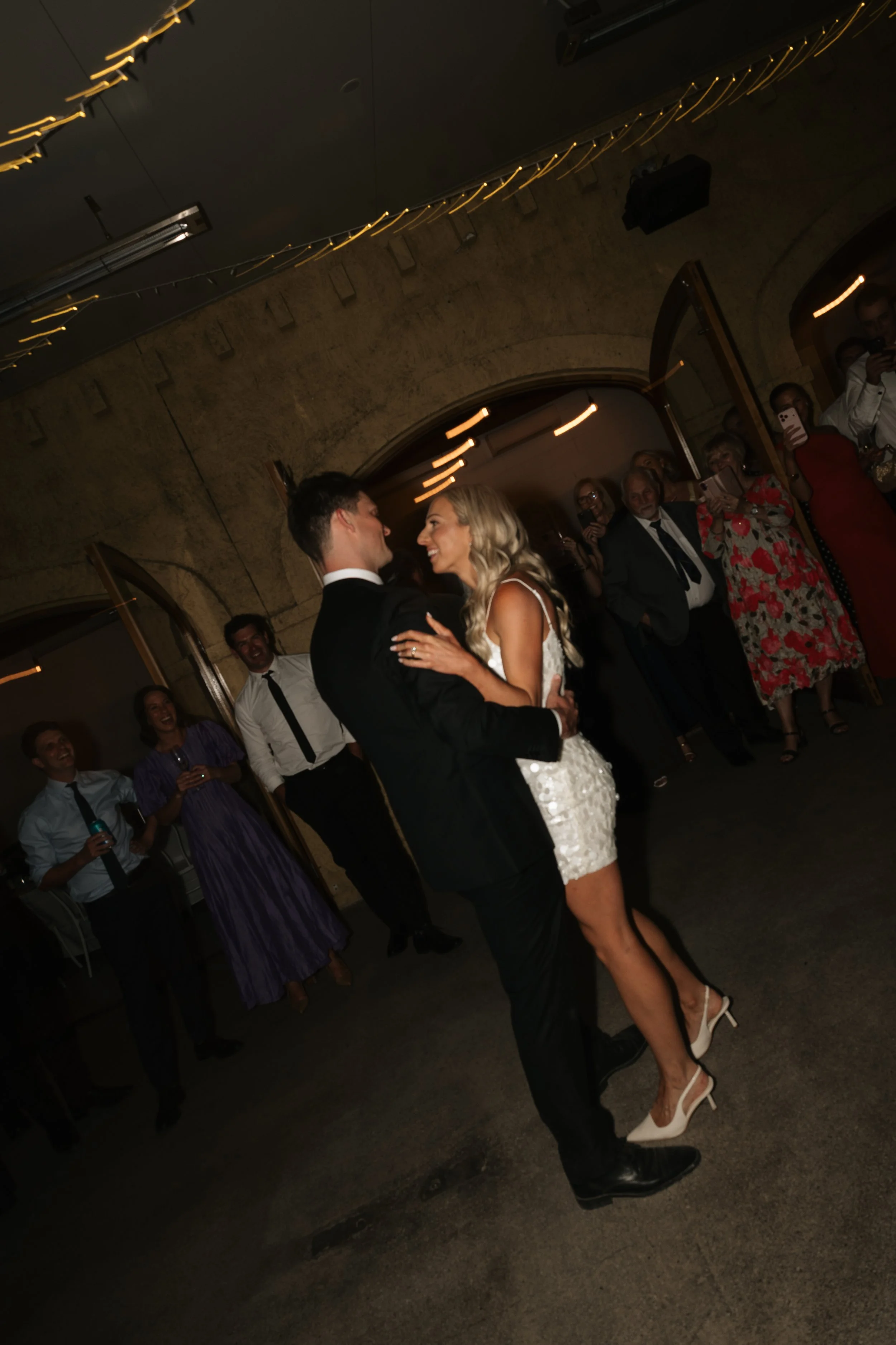 A couple, dressed in formal wedding attire, sharing their first dance at a wedding reception, with guests watching and taking photos in the background, under string lights hanging from the ceiling.