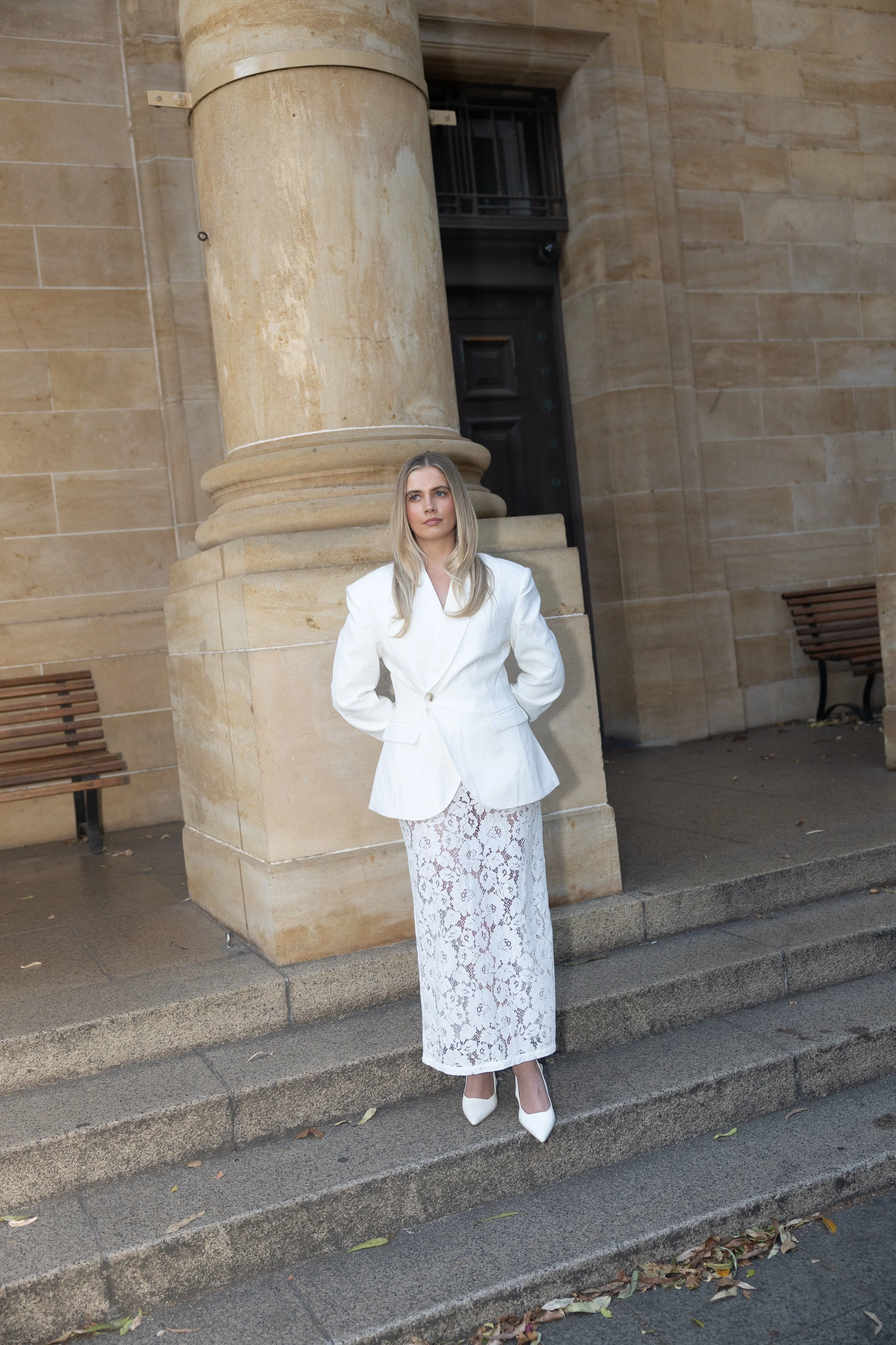 A woman in a white blazer and lace skirt standing on steps in front of a stone building with large columns.