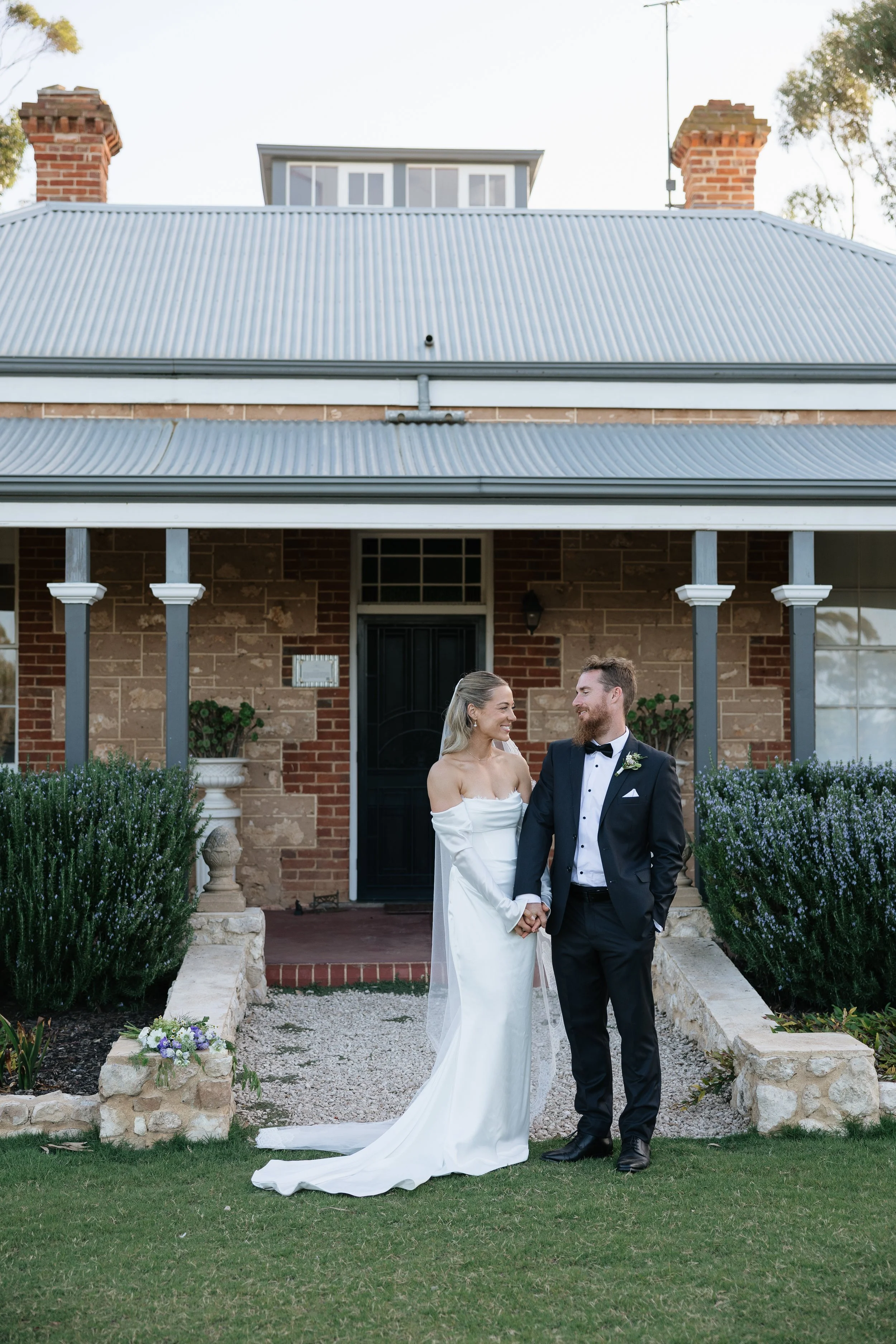 A bride and groom holding hands and smiling at each other in front of a brick house with a metal roof, greenery, and purple flowers. Woodburn Homestead Estate