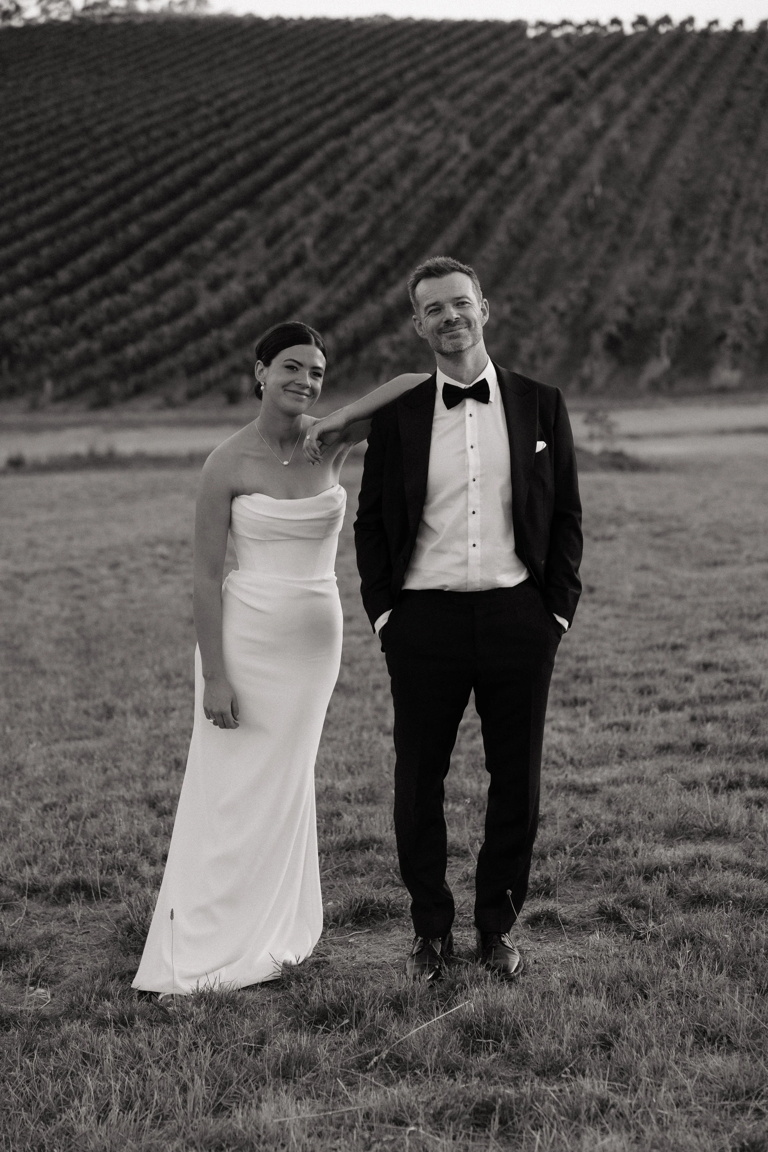 A bride in a strapless white wedding dress and a groom in a black tuxedo standing outdoors in a grassy field with rolling hills in the background.