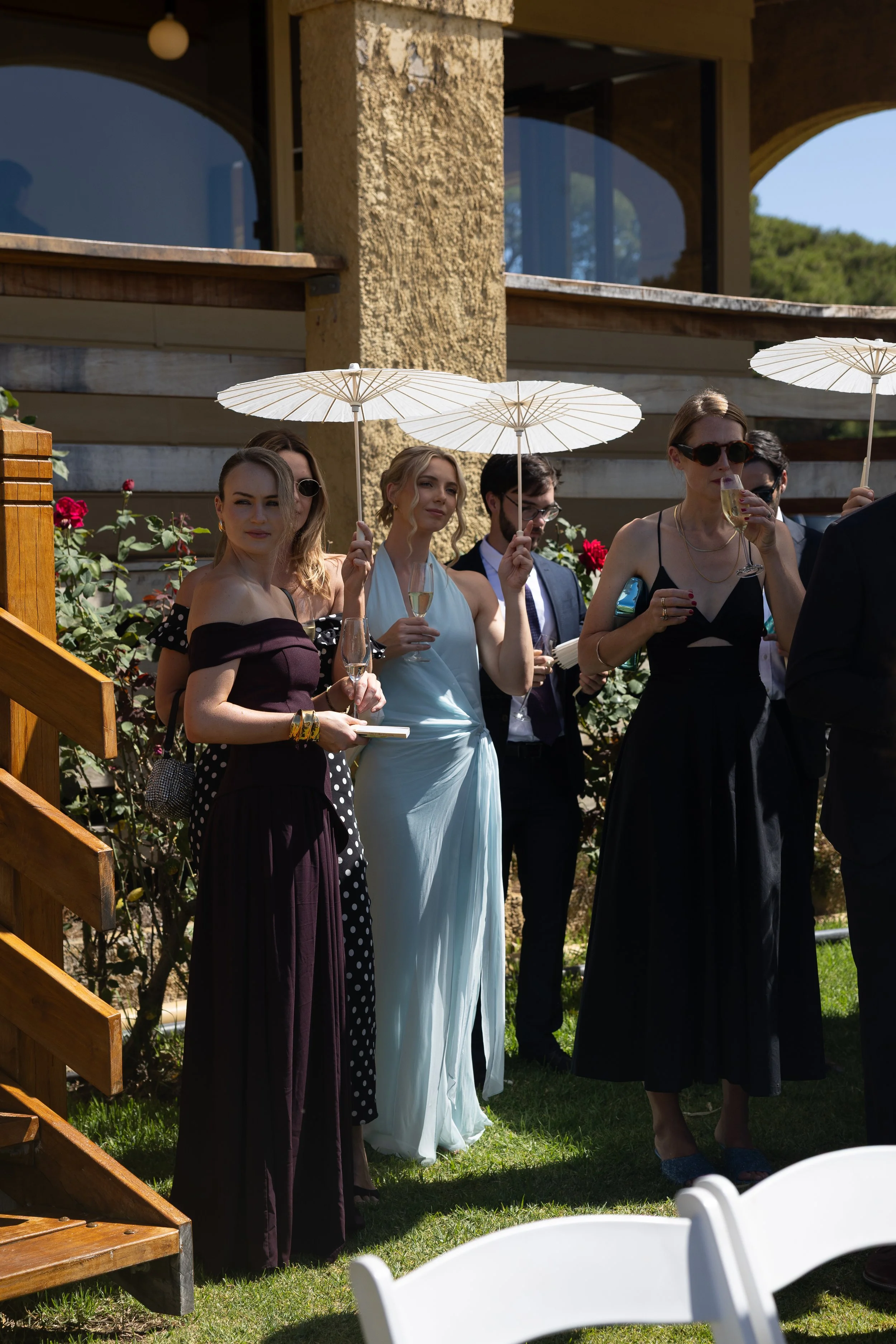 Group of people dressed in formal attire holding drinks and umbrellas at an outdoor social event.
