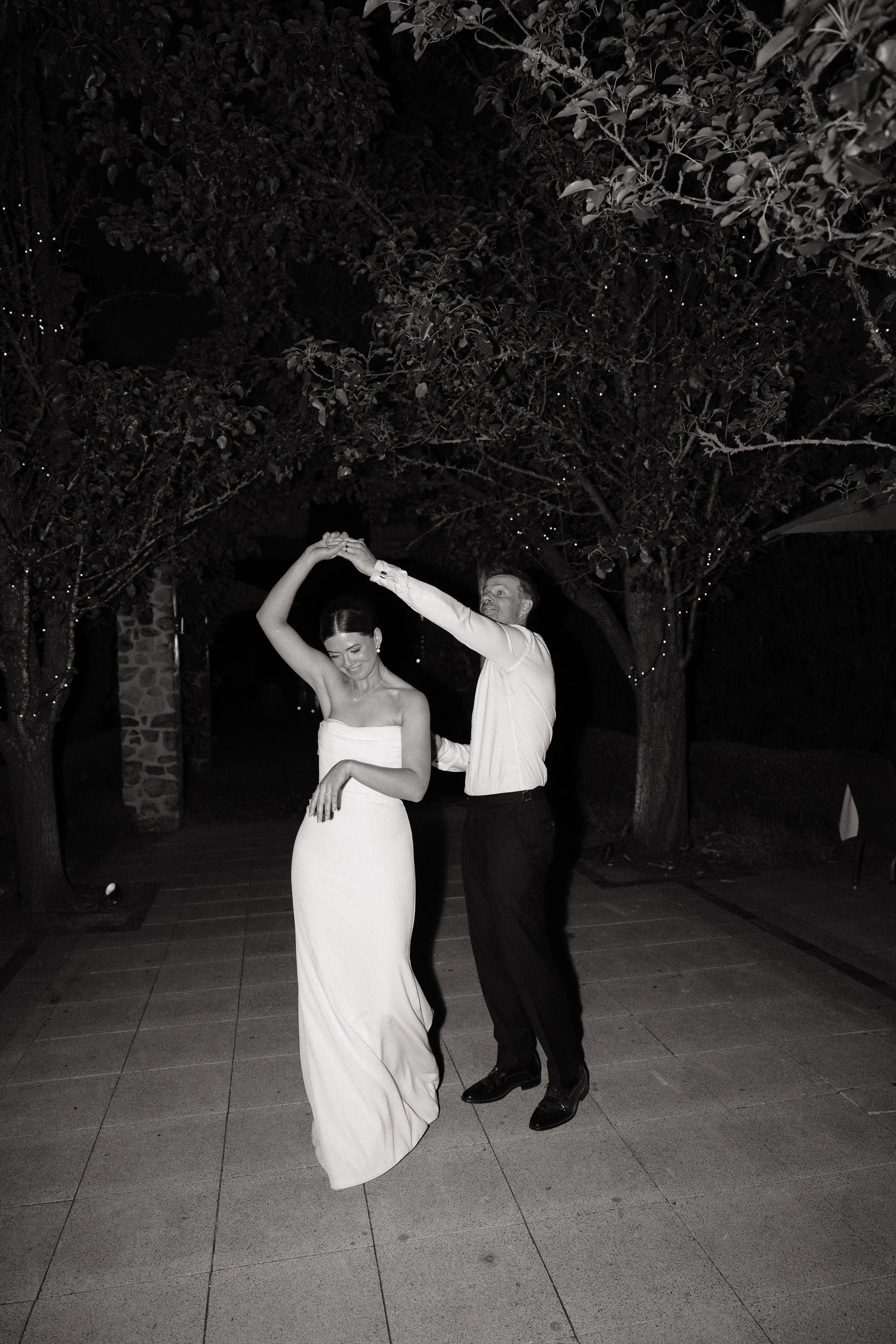 A couple dancing outdoors at night, with trees and string lights in the background, in black and white.