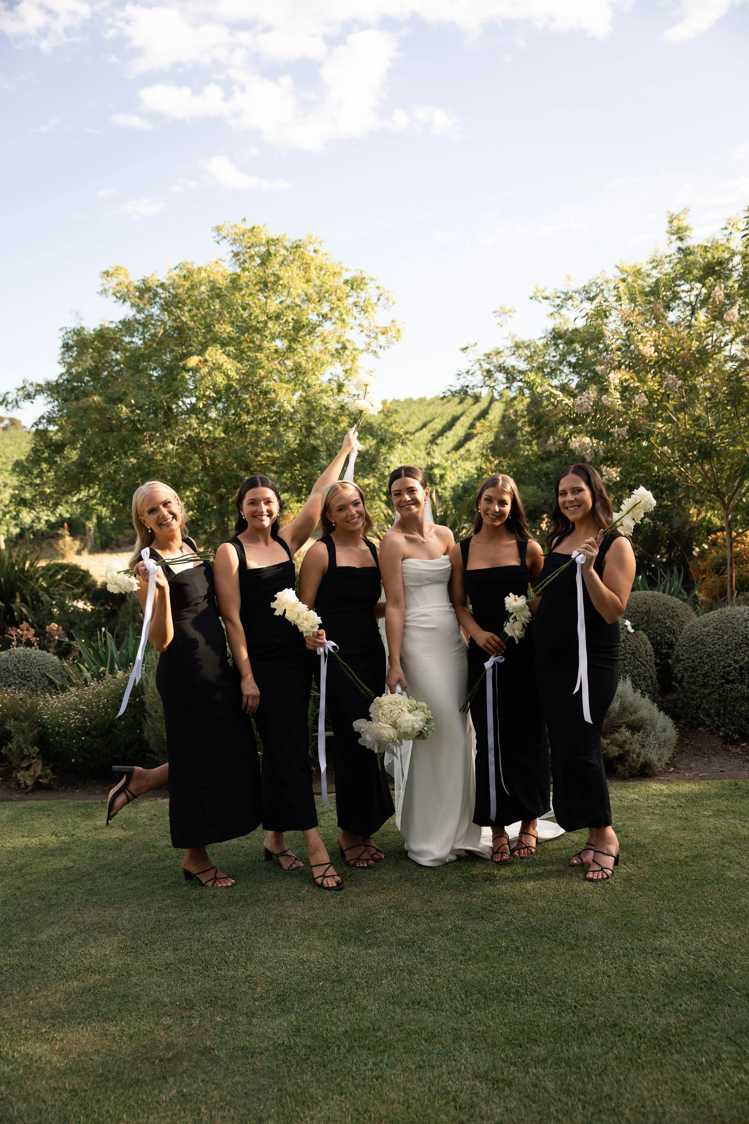 A bride and five bridesmaids in black dresses standing outdoors on grass, holding white flowers, with a garden and trees in the background, under a partly cloudy sky.