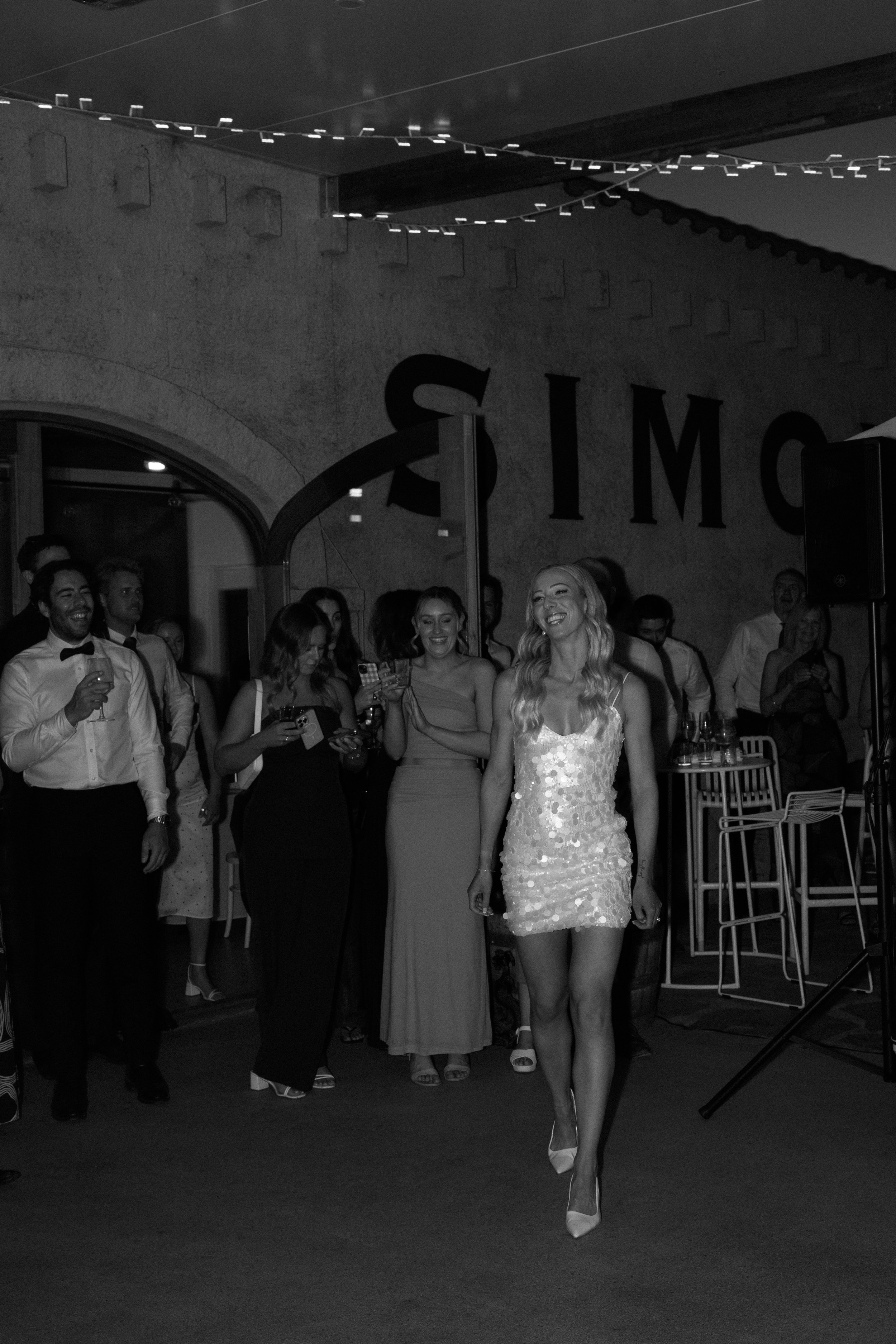 A woman in a sequin dress walking in front of a group of people at a party or celebration, with some holding drinks and taking photos, under a ceiling decorated with string lights.