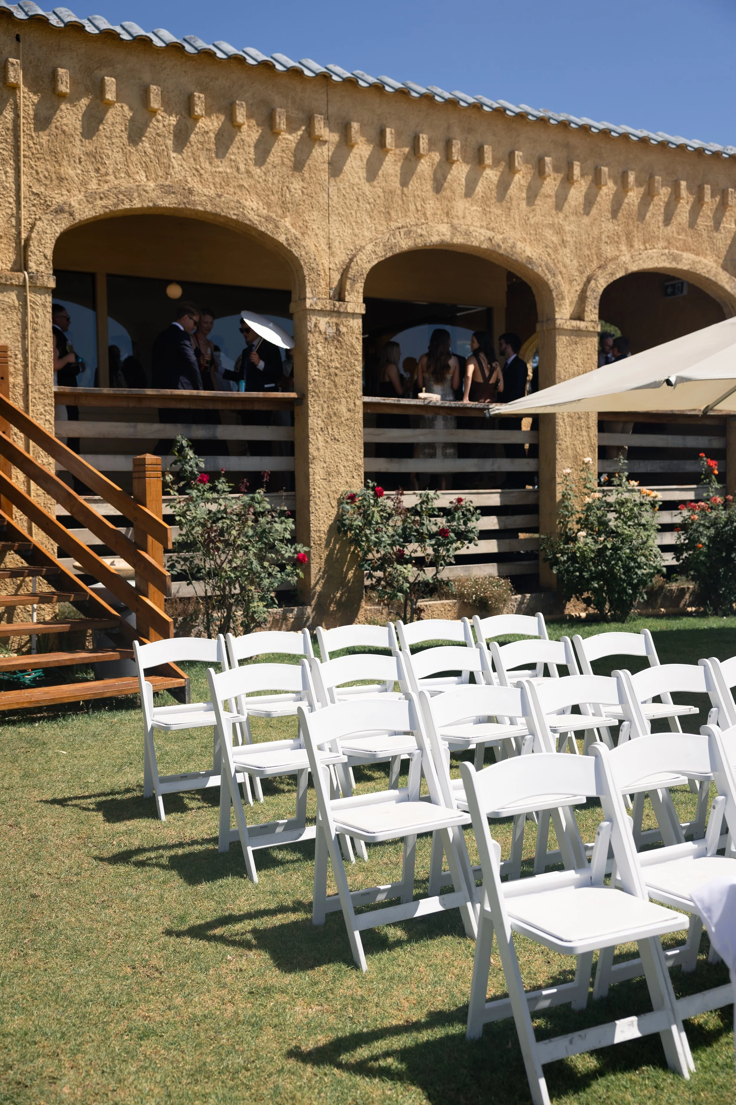 Outdoor wedding or event setup on a lawn with white folding chairs arranged for guests, a building with stone walls, arched openings, and wooden stairs, and guests mingling under an umbrella.