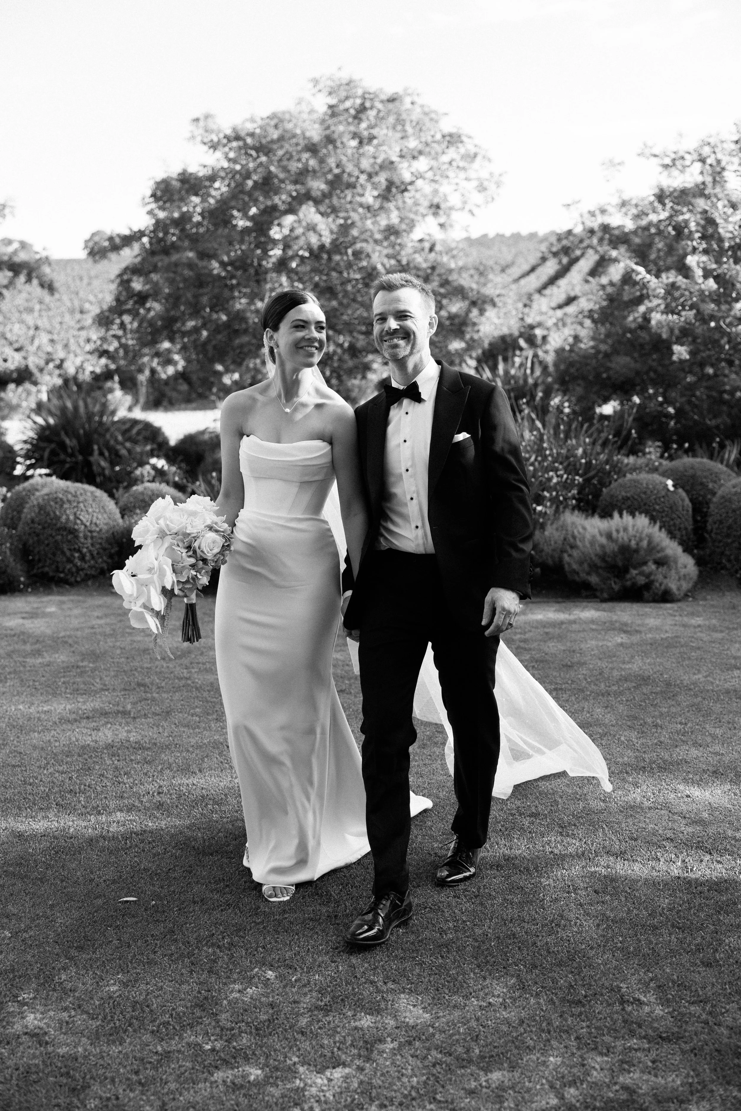 A black and white photograph of a bride and groom walking outdoors. The bride is wearing a strapless wedding gown and holding a bouquet of flowers, while the groom is dressed in a tuxedo with a bow tie. They are smiling and looking at each other, sur