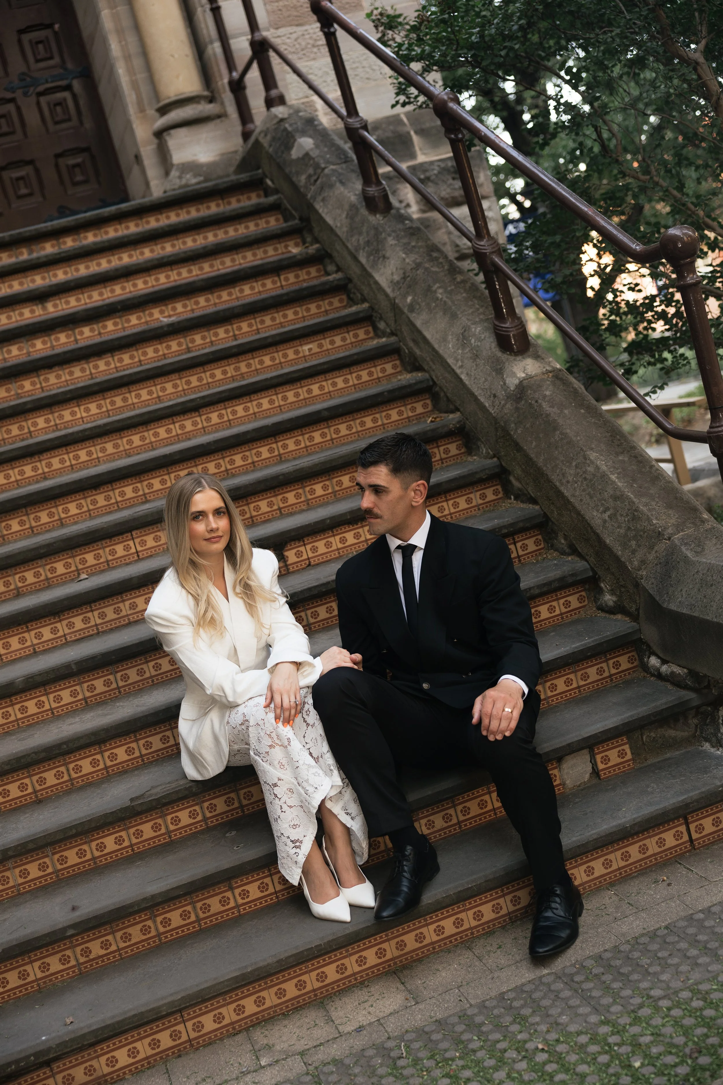A man and woman sitting on outdoor stairs, holding hands. The woman wears a white blazer, lace pants, and white heels; the man wears a black suit and tie. They sit on decorative tiled stairs with a brown railing and greenery in the background.