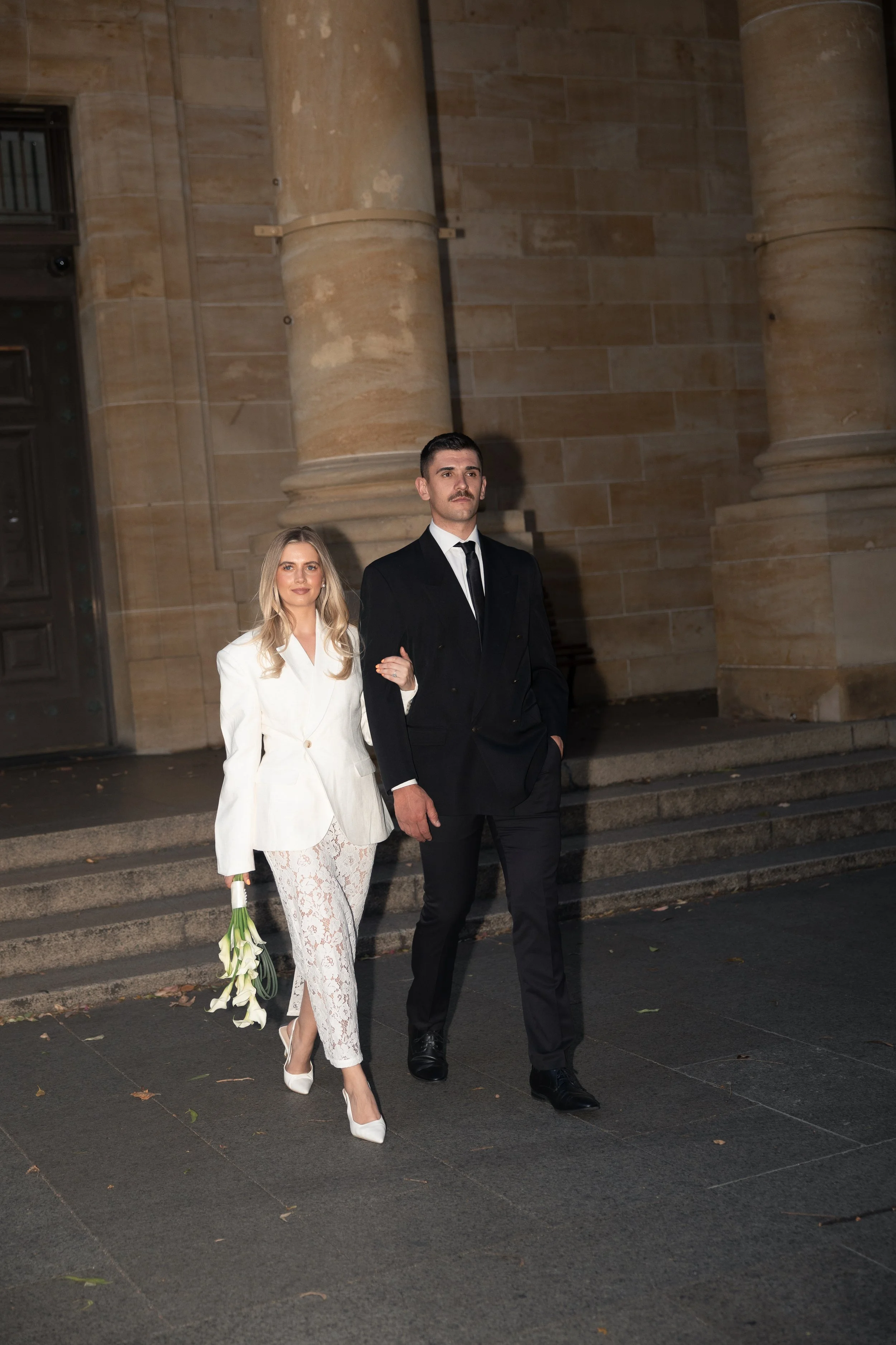 A woman in a white blazer and lace pants holding white calla lilies walking arm-in-arm with a man in a black suit, outside a stone building with large columns.