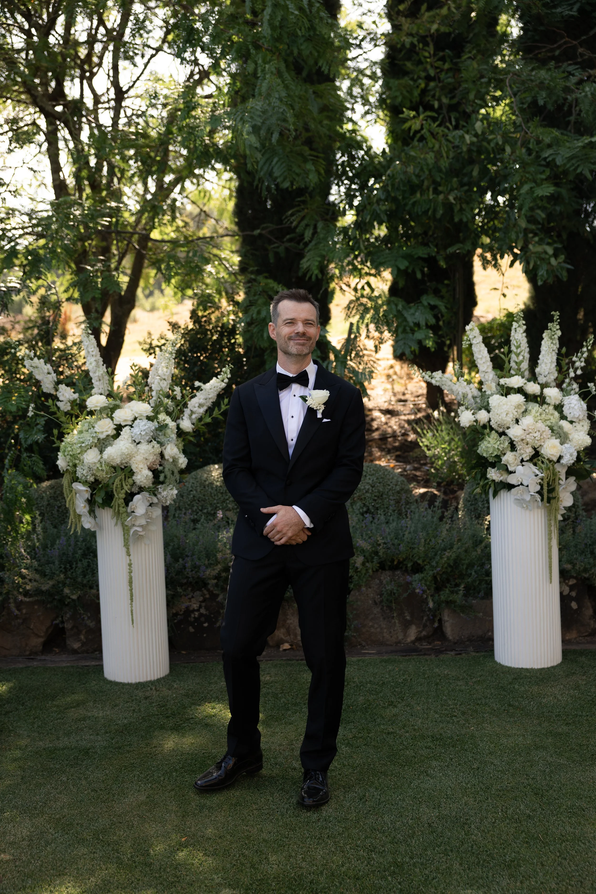 A man dressed in a tuxedo standing outdoors in front of two large floral arrangements, with trees and greenery in the background.