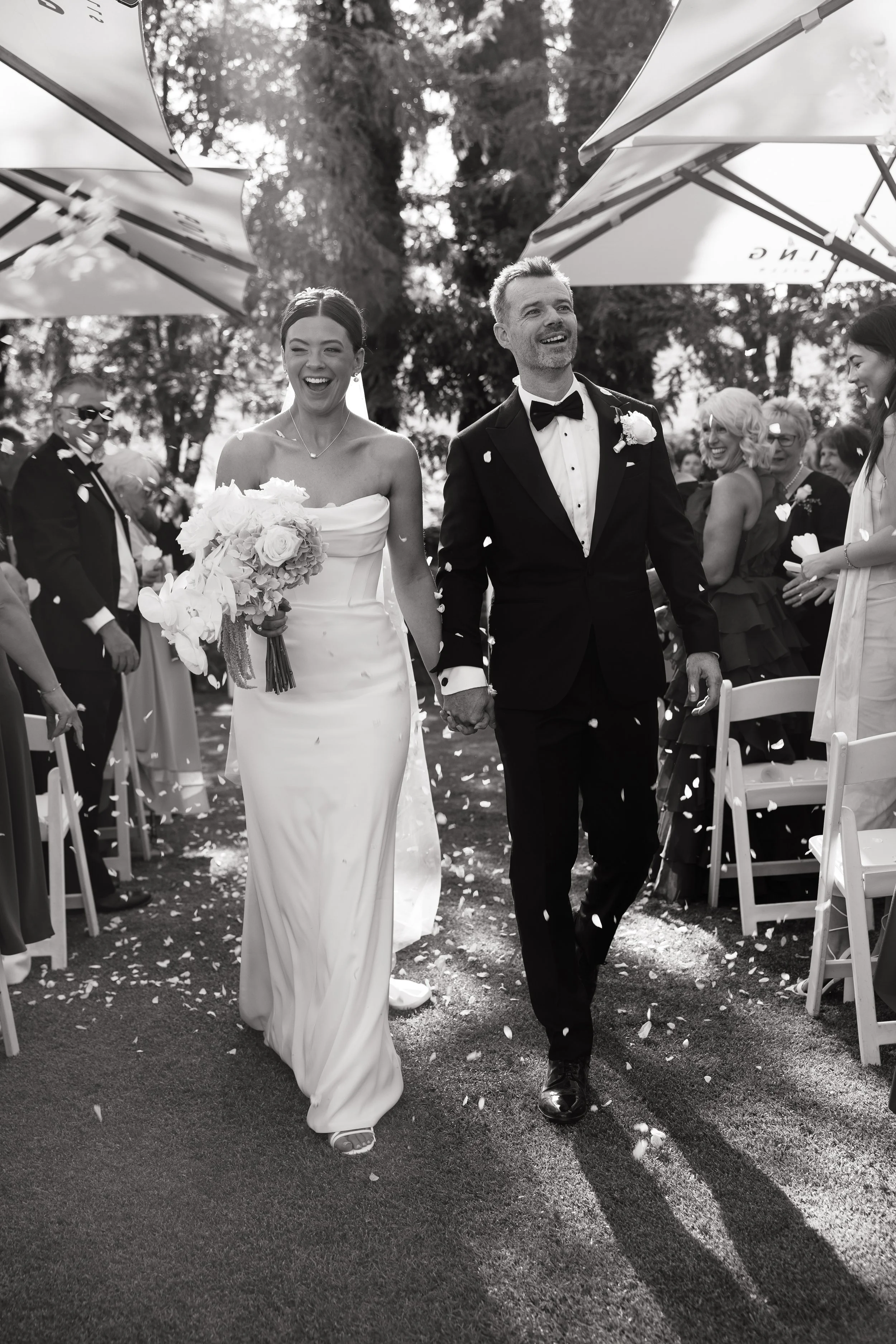 Black and white photo of a bride and groom walking down the aisle. The bride wears a strapless wedding gown and holds a bouquet. The groom wears a tuxedo with a bow tie. Guests are on either side, smiling and celebrating as confetti is thrown.