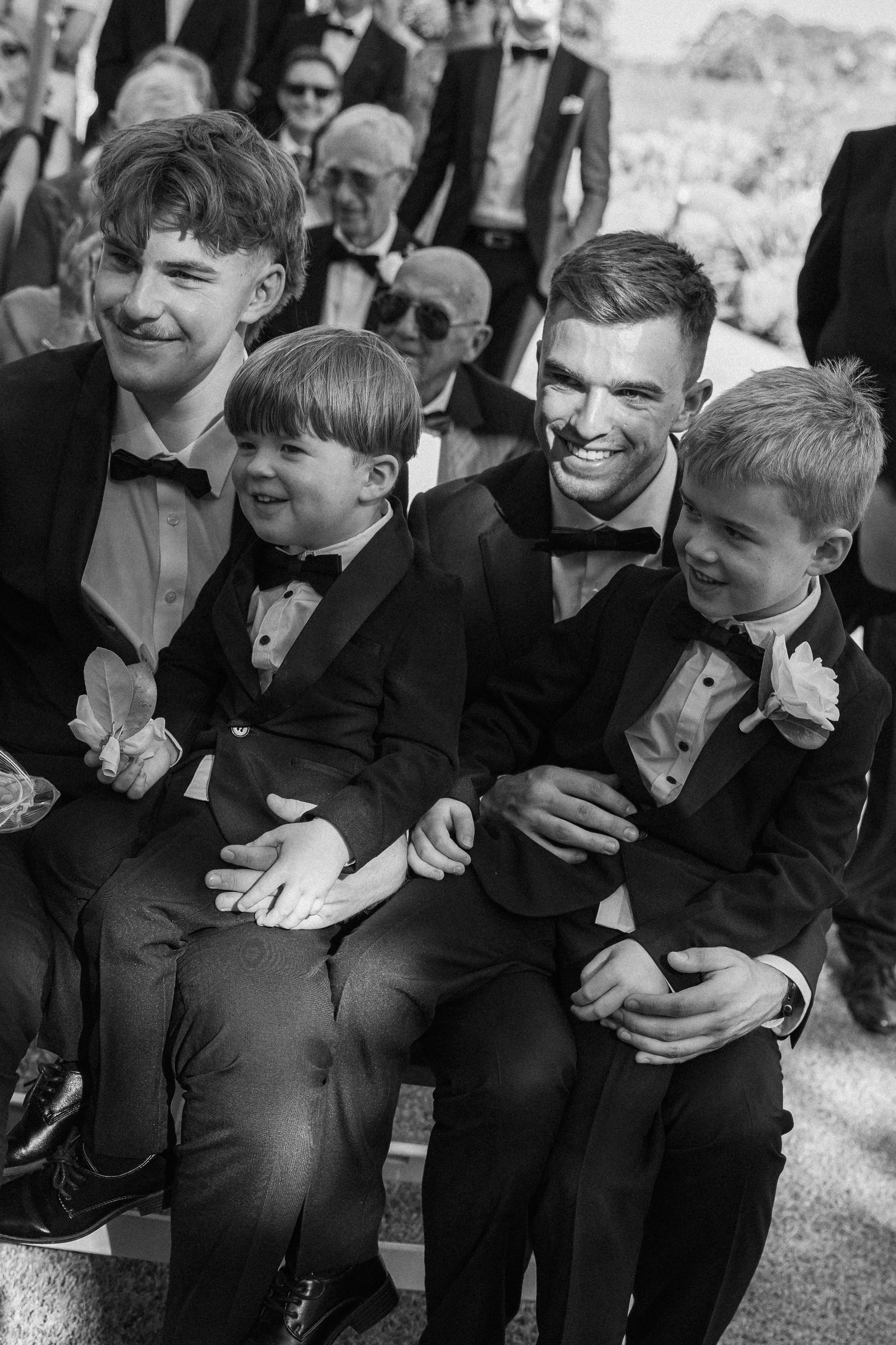 A group of four people, two young boys and two young men, dressed in tuxedos with bow ties, sitting close together at a formal event, smiling and enjoying the moment. In the background, other people in formal attire are visible, some wearing sunglass