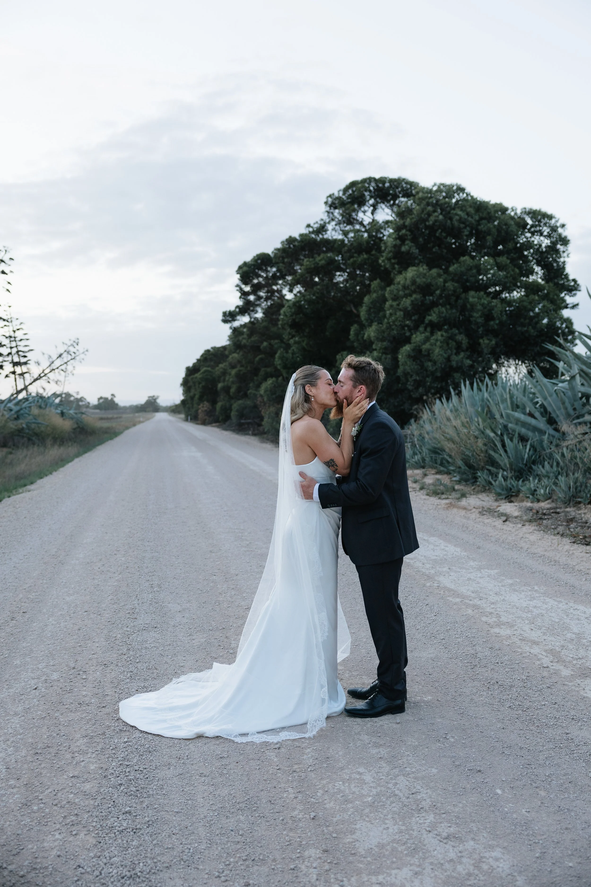 A bride and groom stand close together on a dirt road, sharing a kiss during their wedding, with trees and cloudy sky in the background. Woodburn Homestead