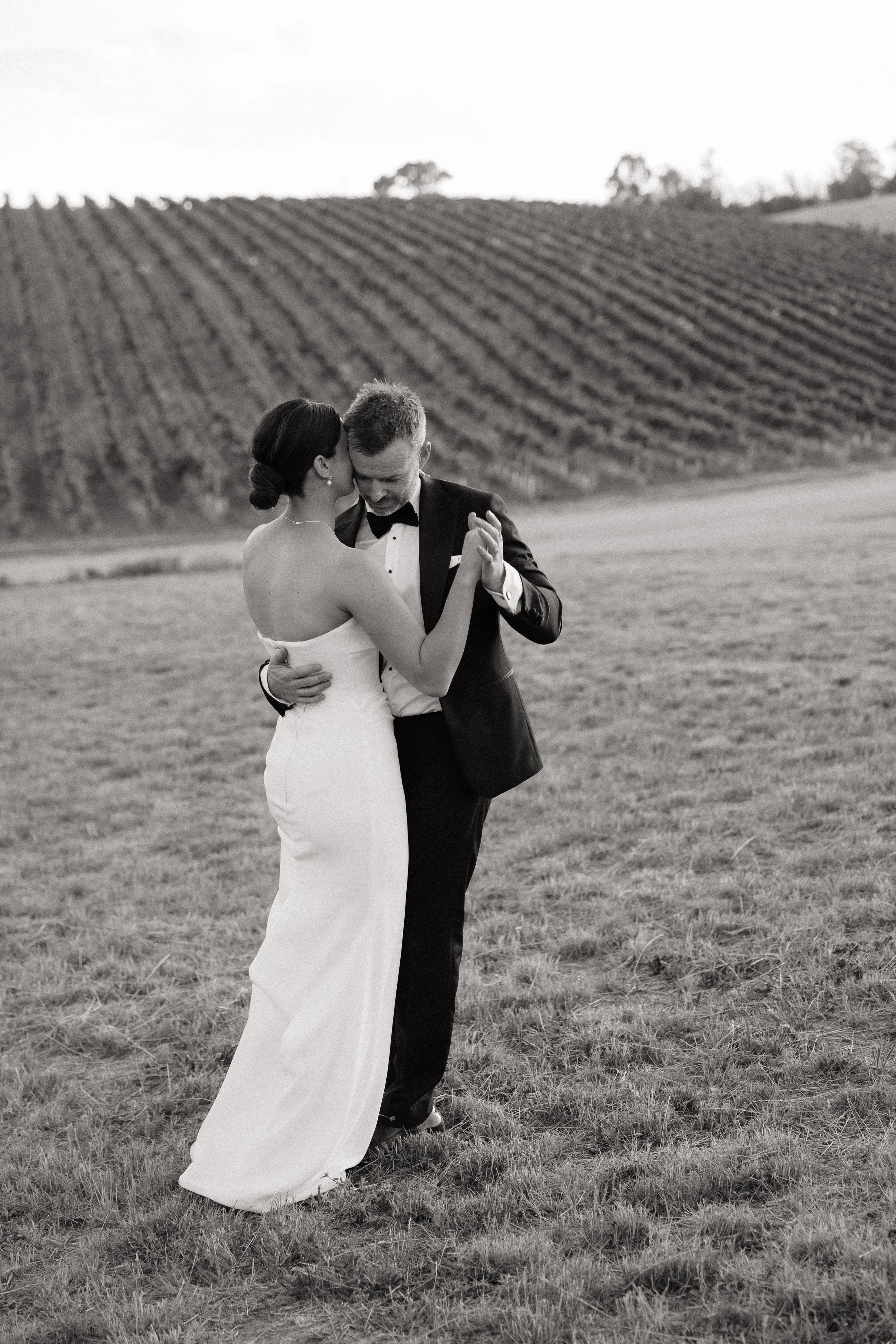 A black and white photo of a bride and groom dancing outdoors on a grassy field with rolling hills and vineyard rows in the background.