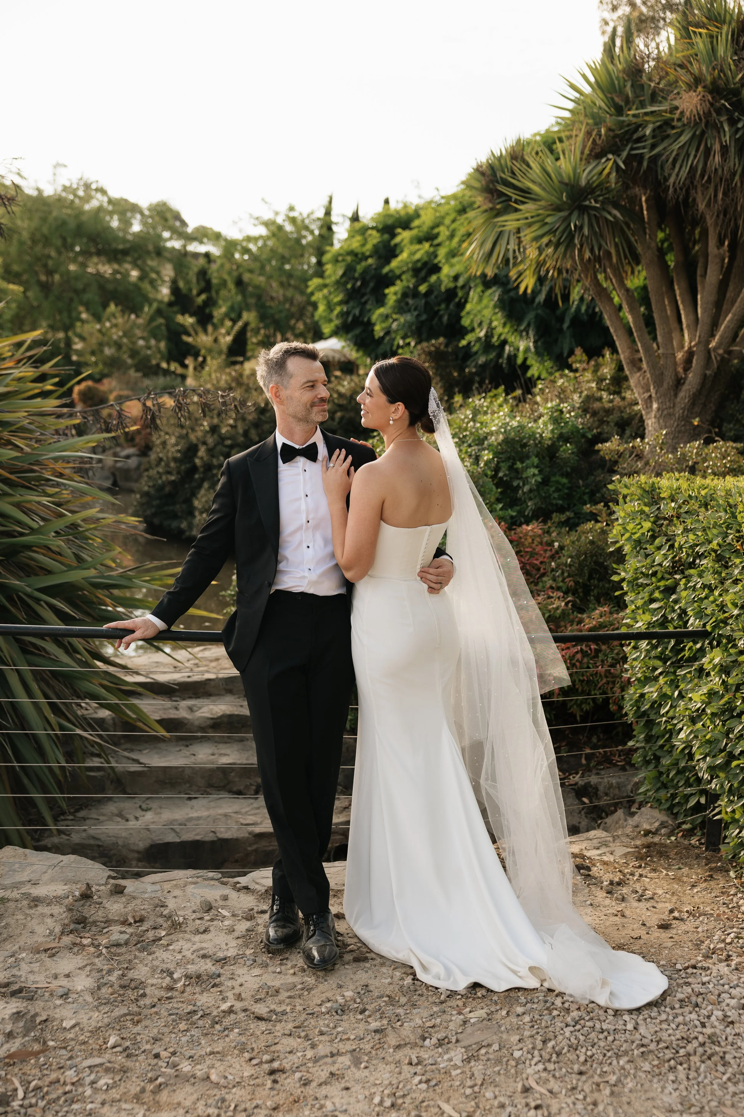 A bride and groom in wedding attire standing outdoors on a dirt path, surrounded by green trees and bushes, with the bride looking at the groom and the groom looking slightly away. Golding Wines Wedding
