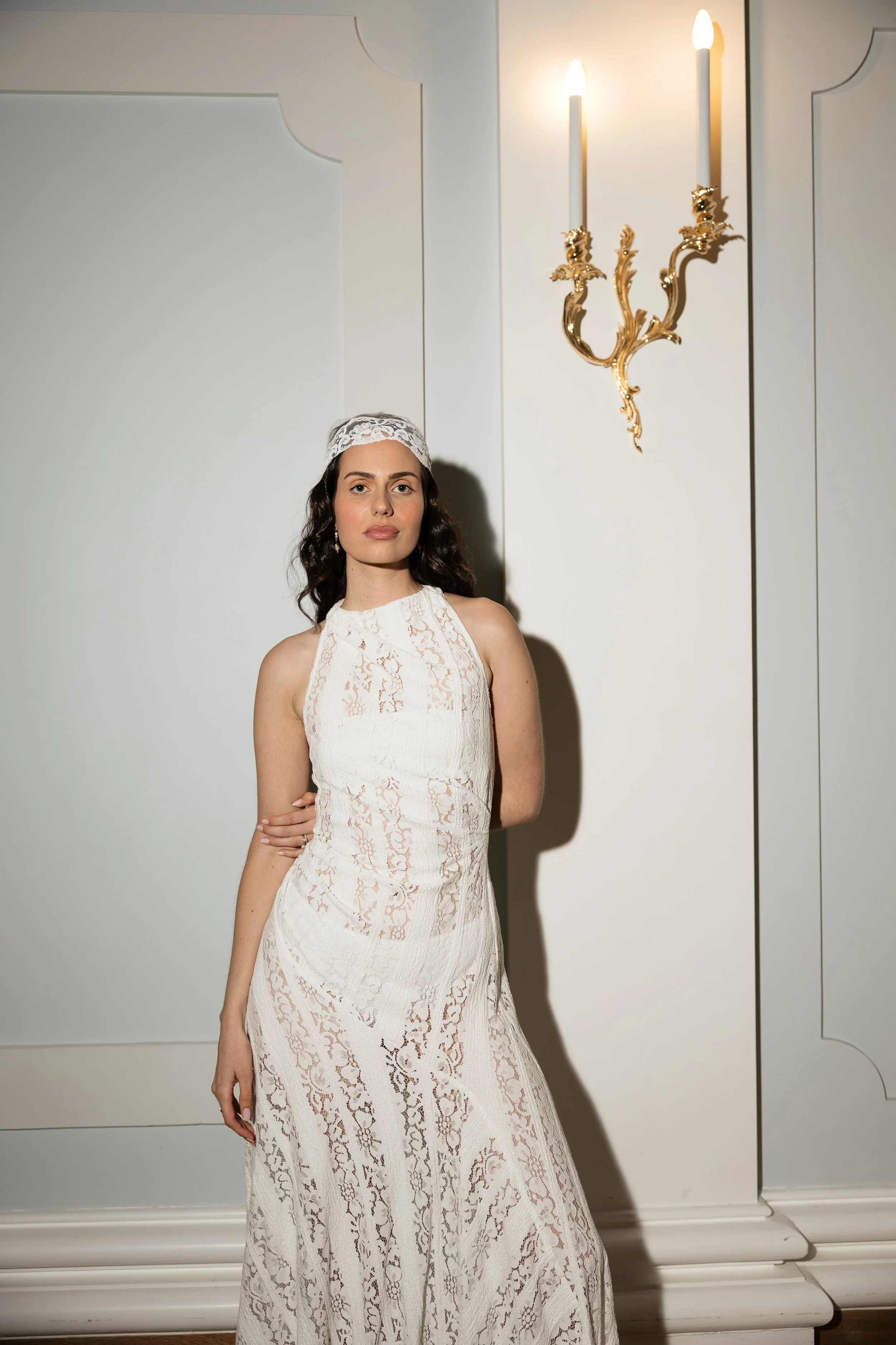 Woman in a white lace dress and head covering standing beside an ornate gold wall sconce