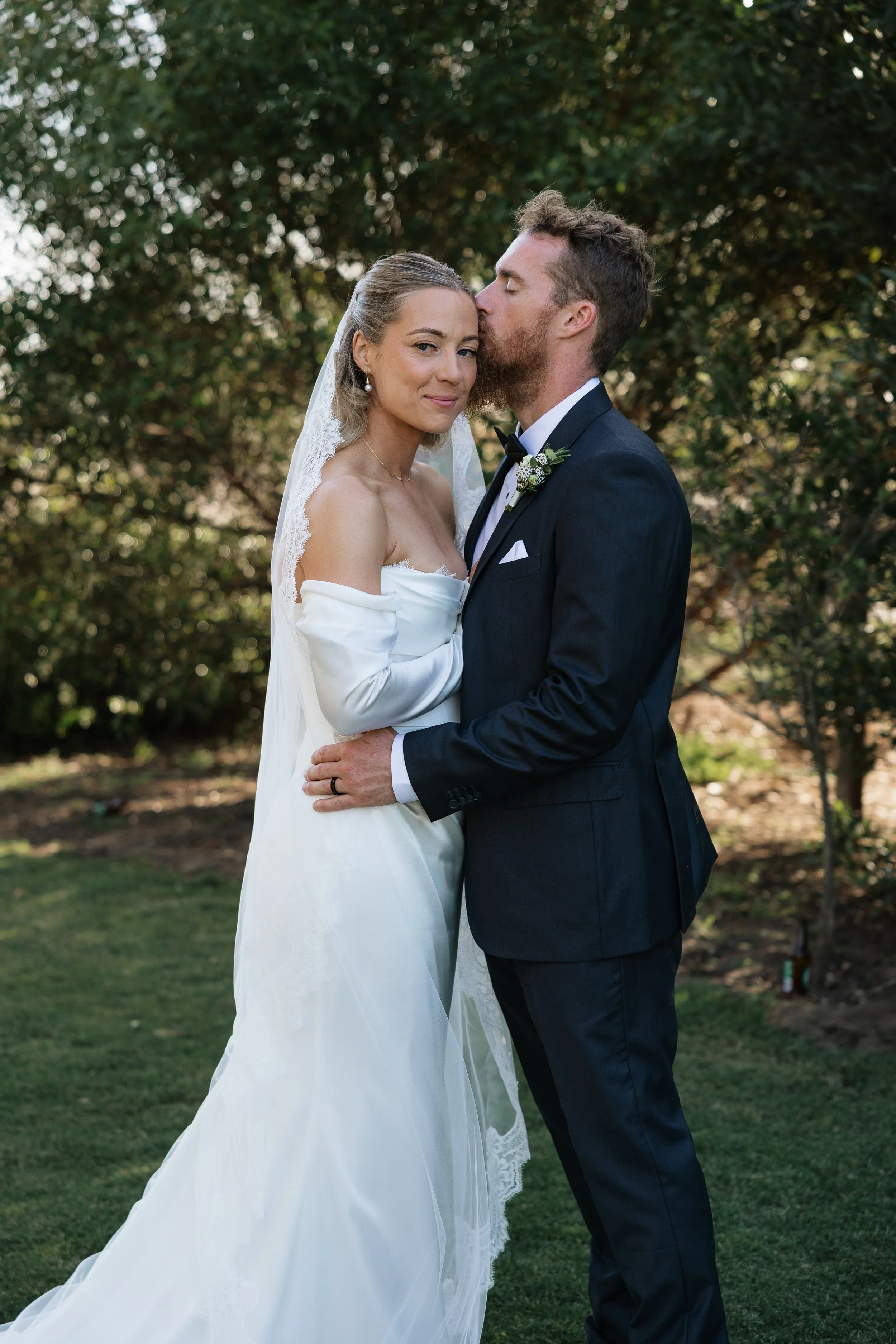 Bride in an off-the-shoulder wedding dress and veil standing outdoors with groom in a black tuxedo, kissing her forehead, against a background of trees. Woodburn Homestead Estate.