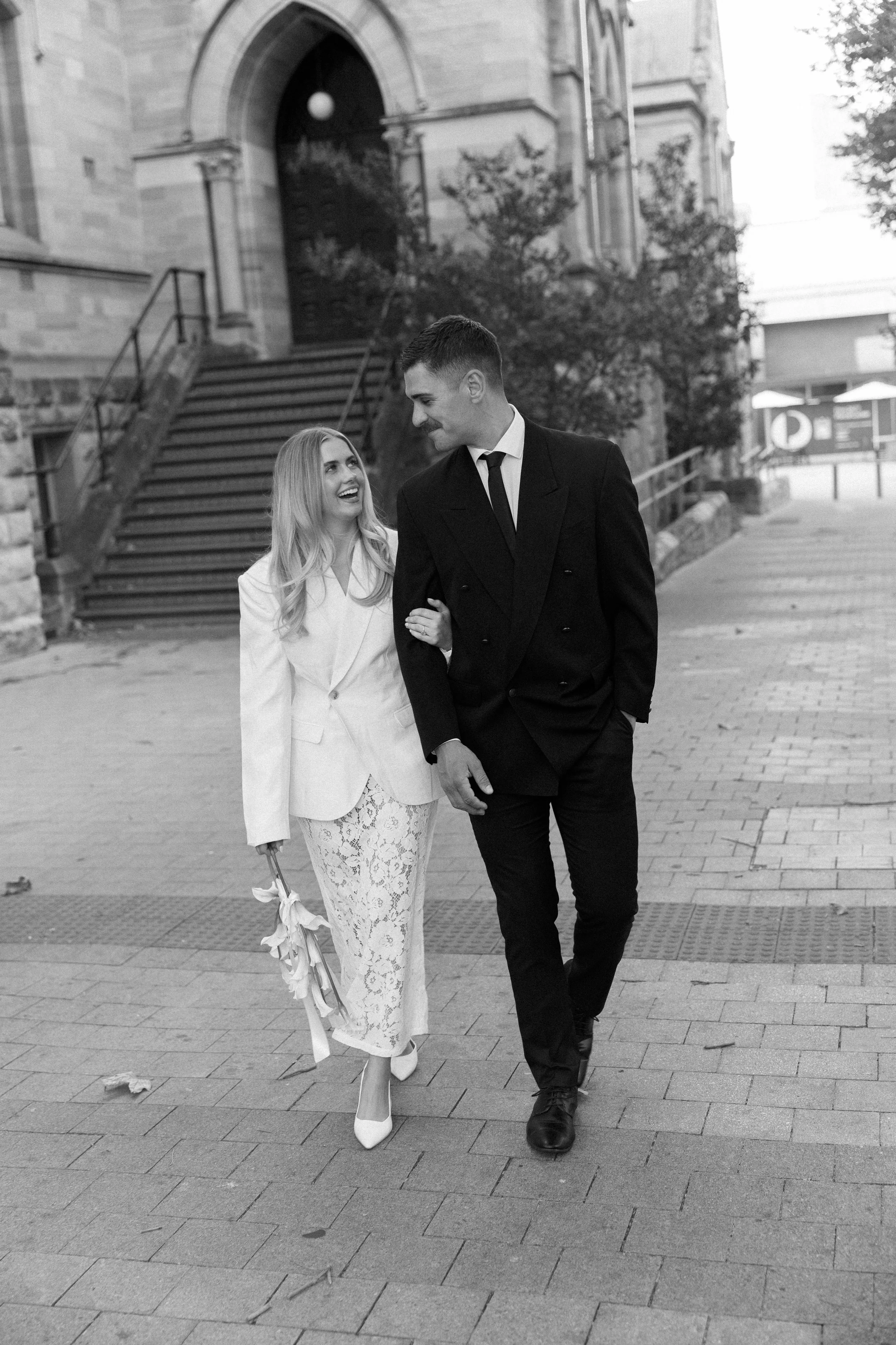 A smiling couple dressed in formal attire walking arm in arm on a city sidewalk, with a historic building in the background.
