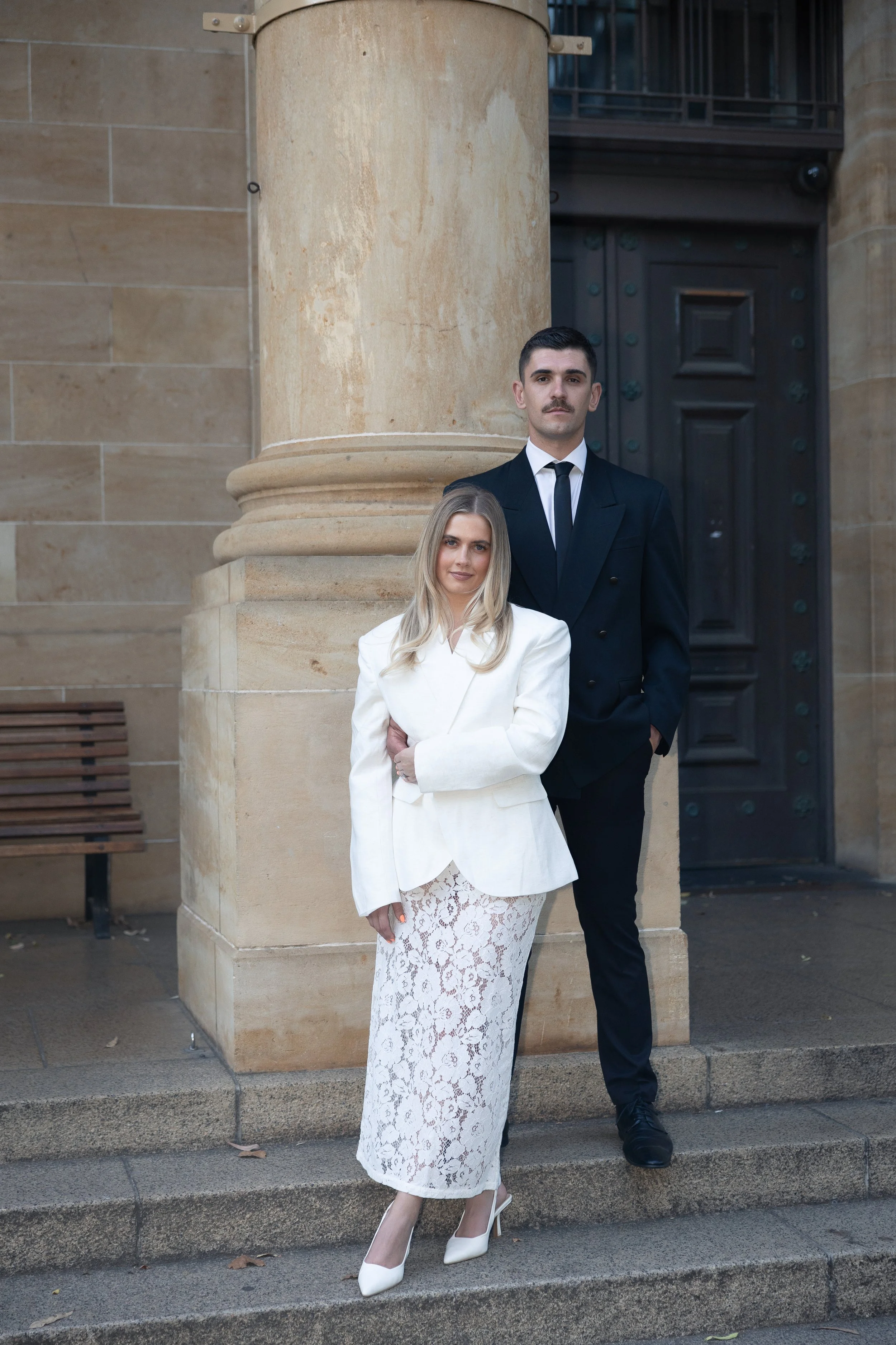 A man and woman dressed in formal attire standing outdoors in front of a large stone building with a dark wooden door.