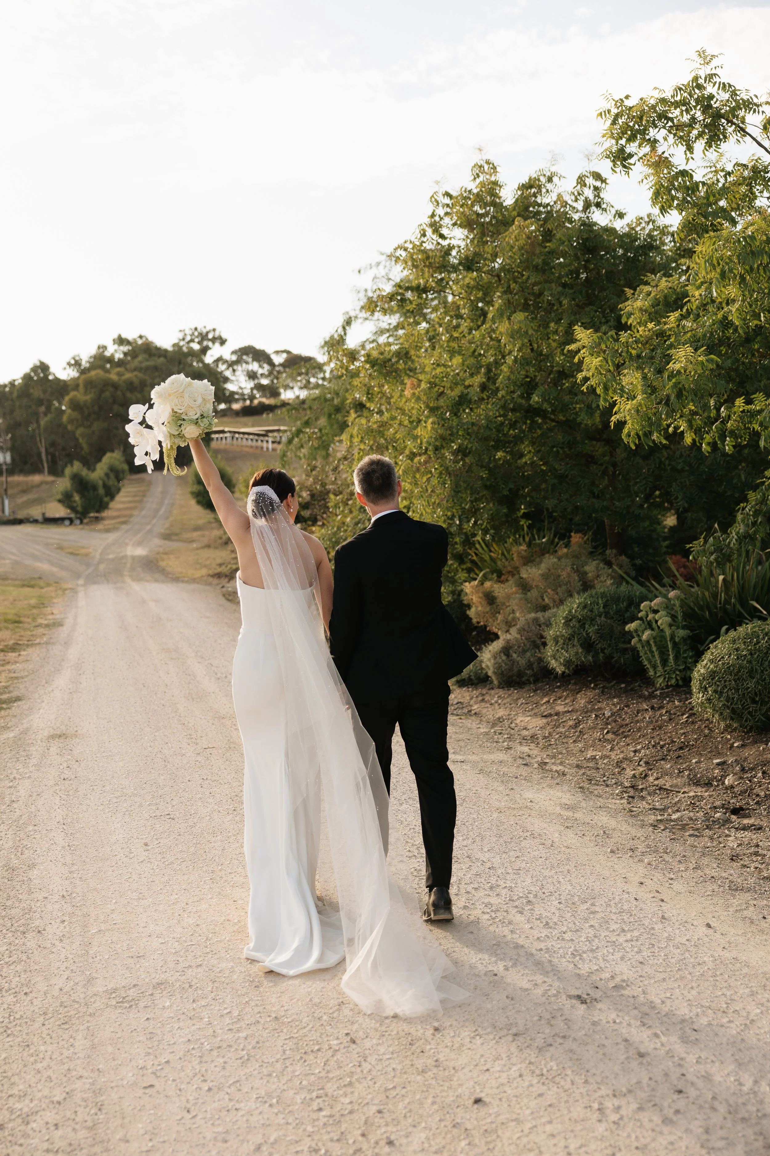 Bride and groom walking down a dirt road during sunset, surrounded by greenery, with the bride holding a bouquet and wearing a white wedding dress and veil. Golding Wines Wedding