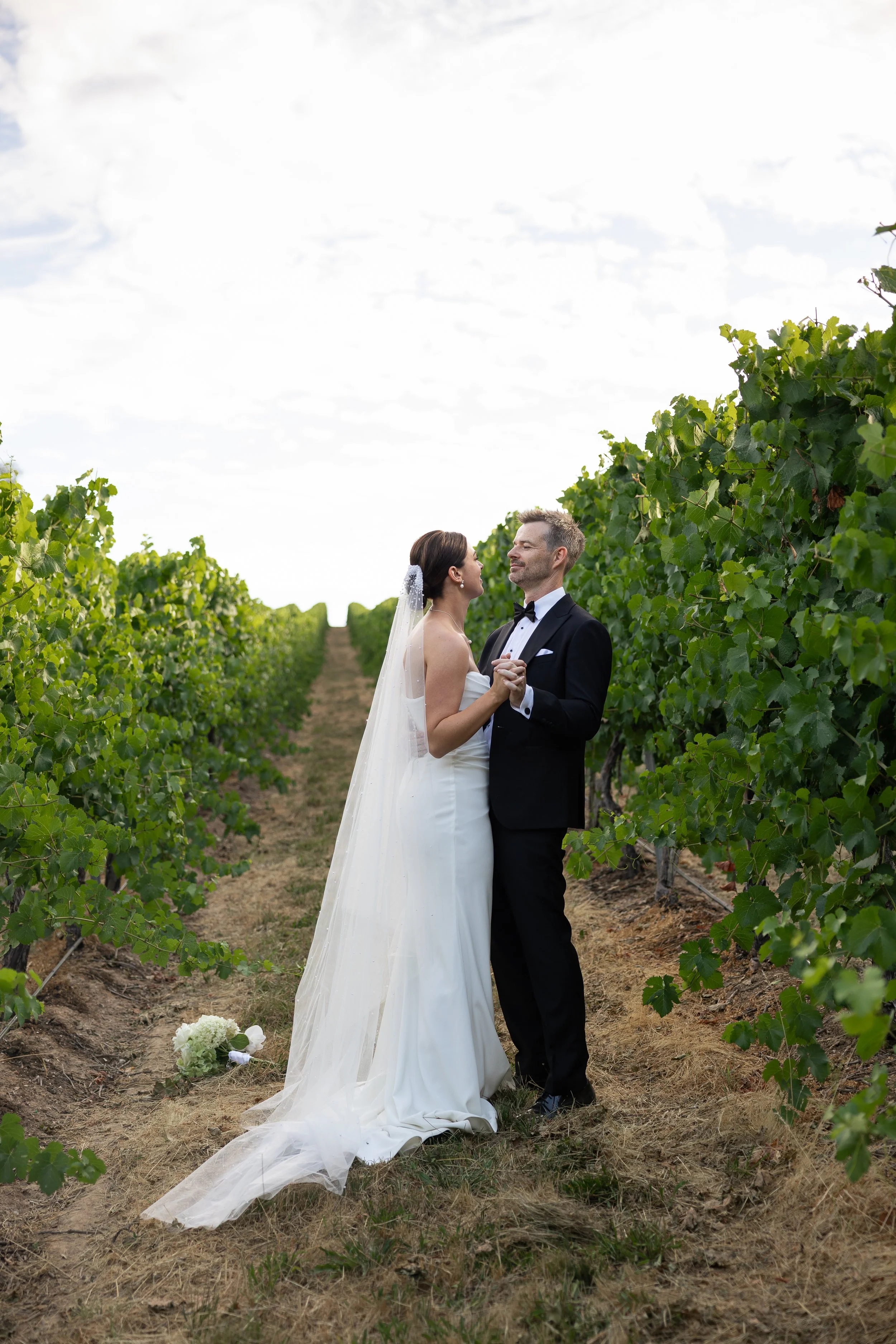 A bride and groom holding hands and looking at each other in a vineyard during their wedding, with bouquet on the ground nearby. Golding Wines Wedding