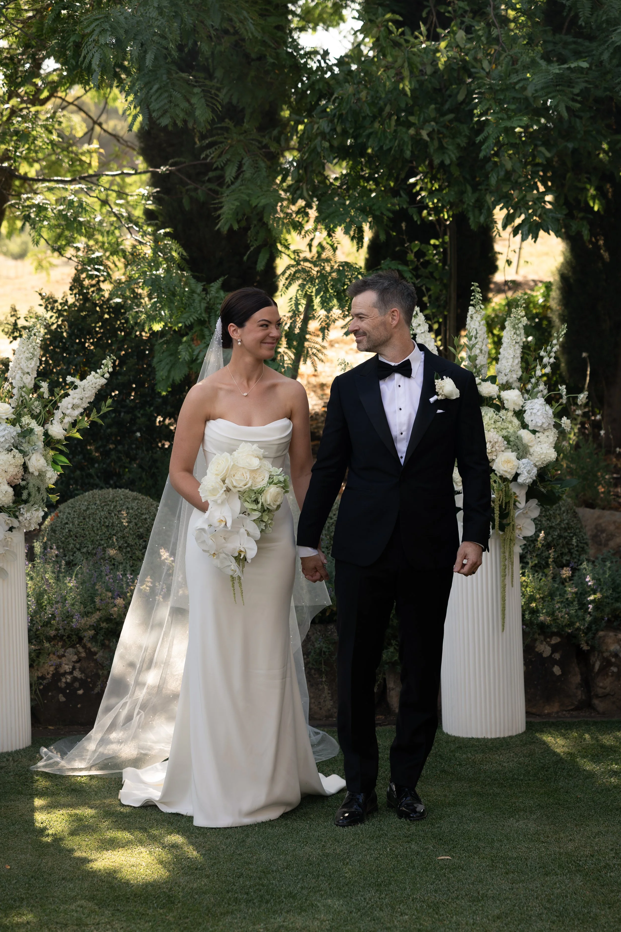 A bride and groom holding hands and smiling at each other during their wedding ceremony outdoors, with white floral arrangements and lush green trees in the background.