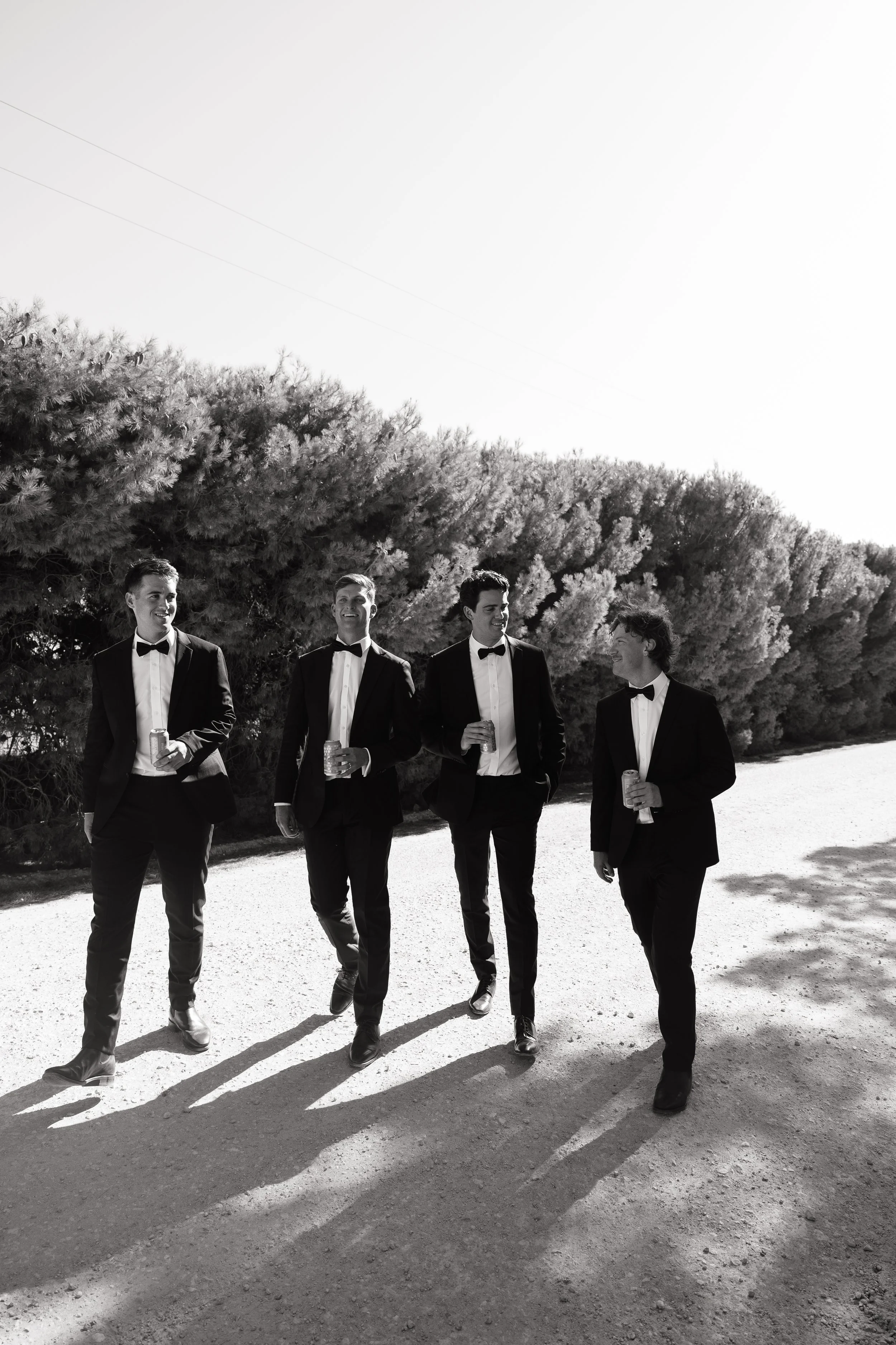 Four men in tuxedos walking outdoors, holding drinks, with shadows cast on the ground, trees in the background, in black and white.