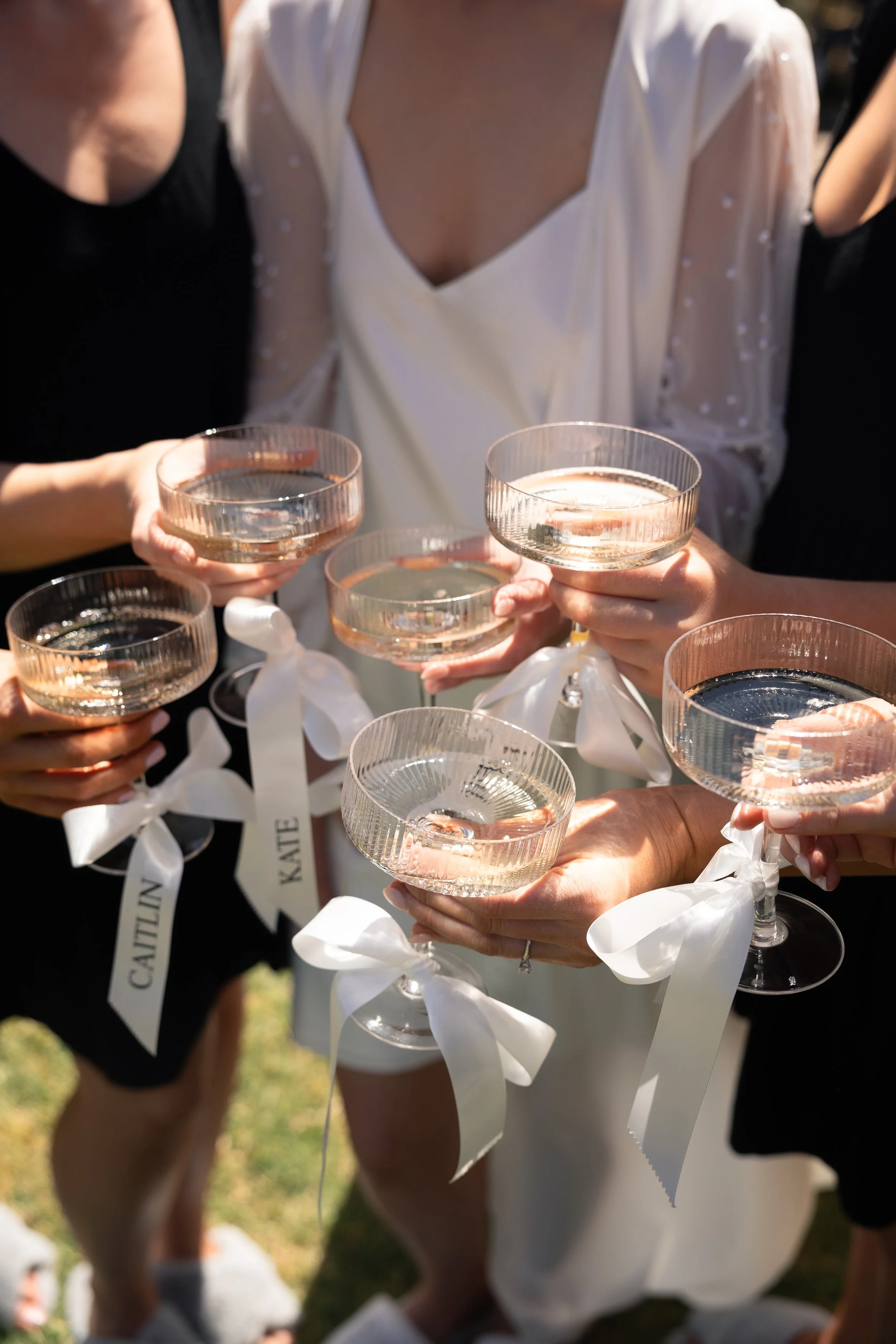 Group of women holding champagne glasses with white ribbons tied around the stems, celebrating outdoors.