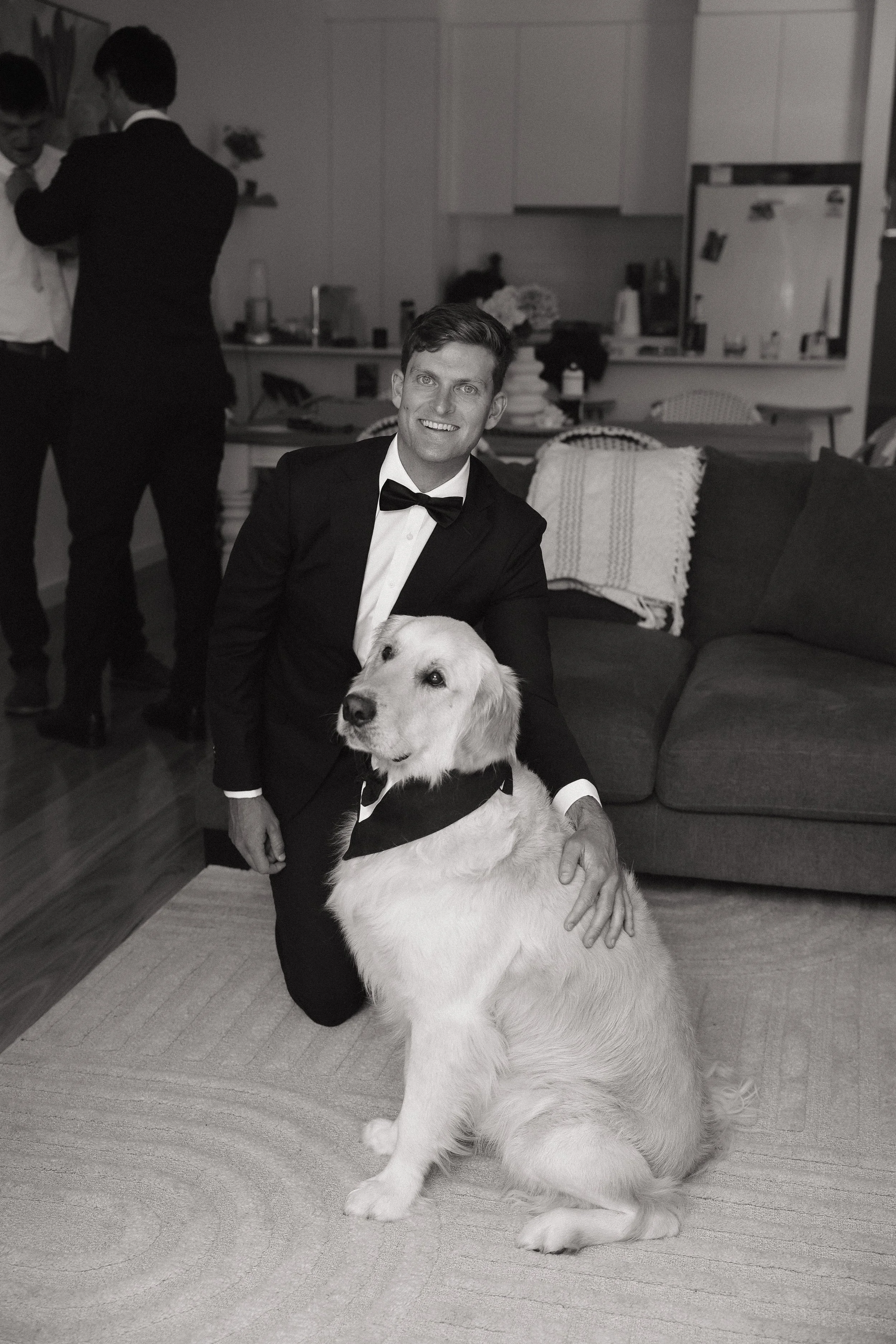 A man in a tuxedo sitting and smiling with a Labrador retriever dog, who is also dressed in a tuxedo, inside a living room with a sofa, pillows, and a kitchen in the background. Two other men are standing and talking in the background. Simon Hackett 