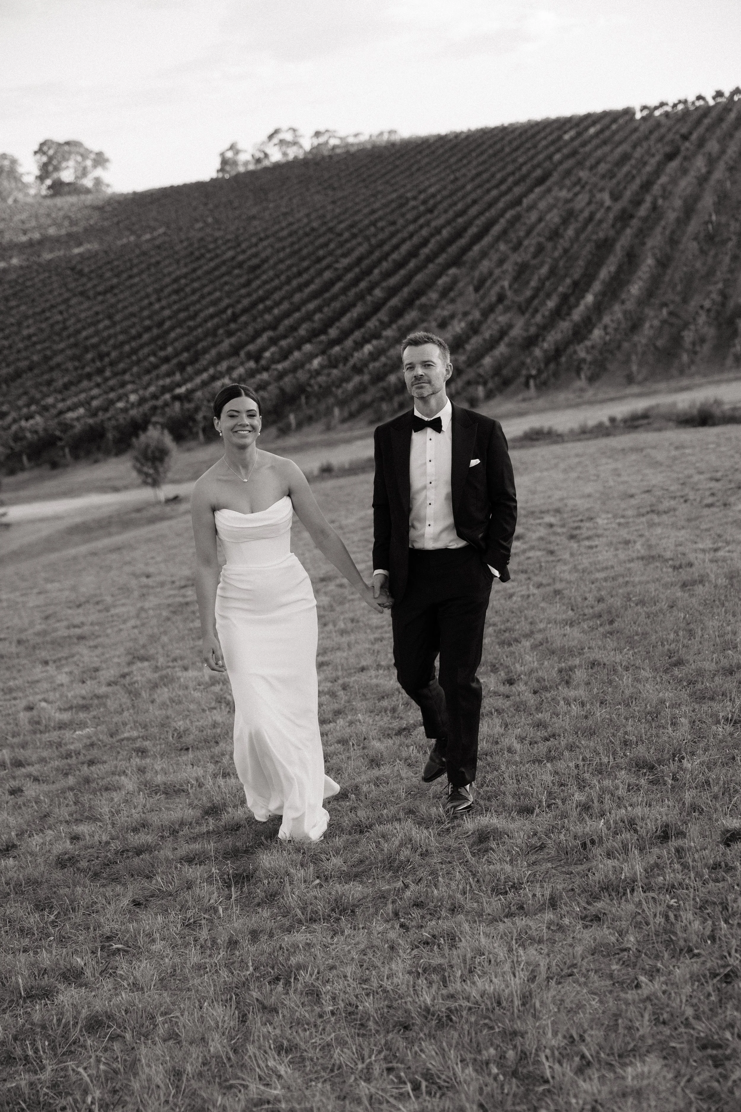 A black-and-white photo of a bride and groom walking hand in hand across a grassy field, with a vineyard on a hillside in the background.