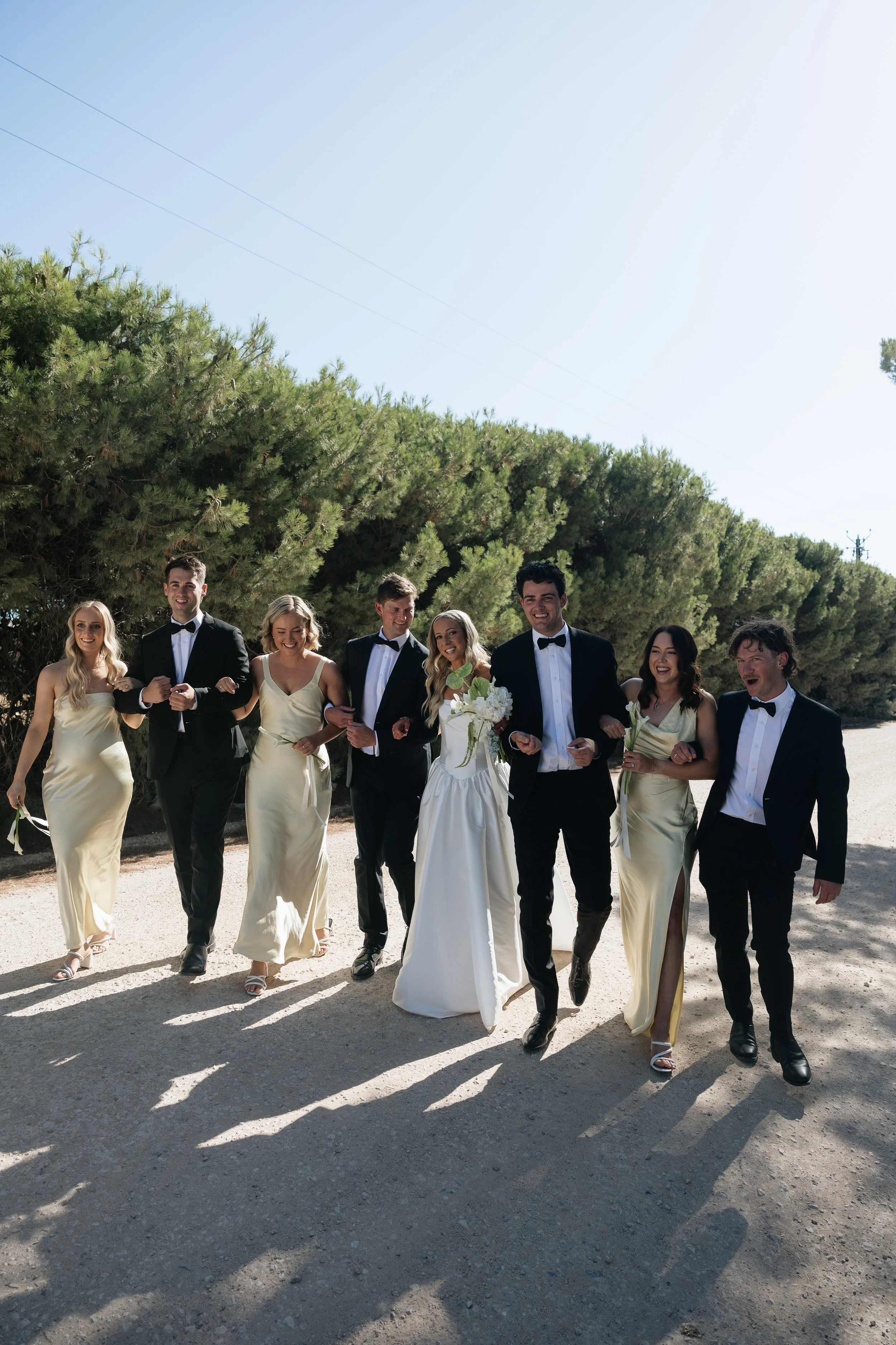 A group of people, dressed in formal attire, walking on a dirt path outdoors with greenery and trees in the background.