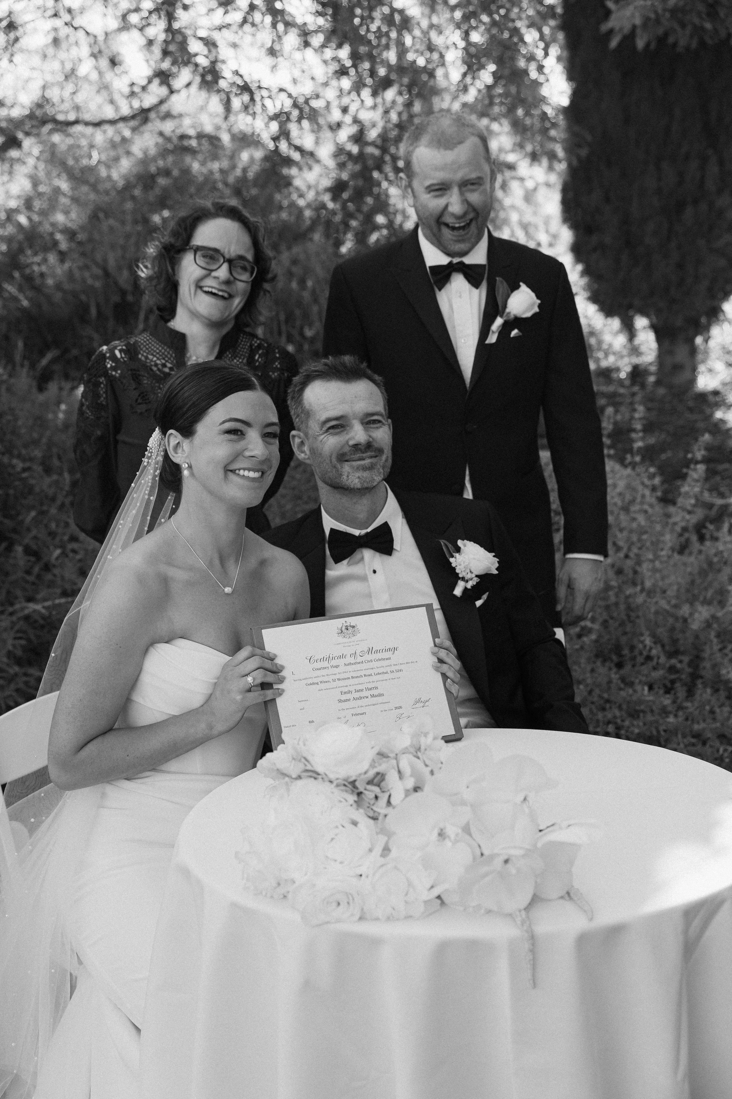 Black and white photo of a wedding celebration outdoors. The bride and groom are sitting at a table with a bouquet, holding a marriage certificate. Two people stand behind them, smiling and dressed formally. The setting is surrounded by trees.