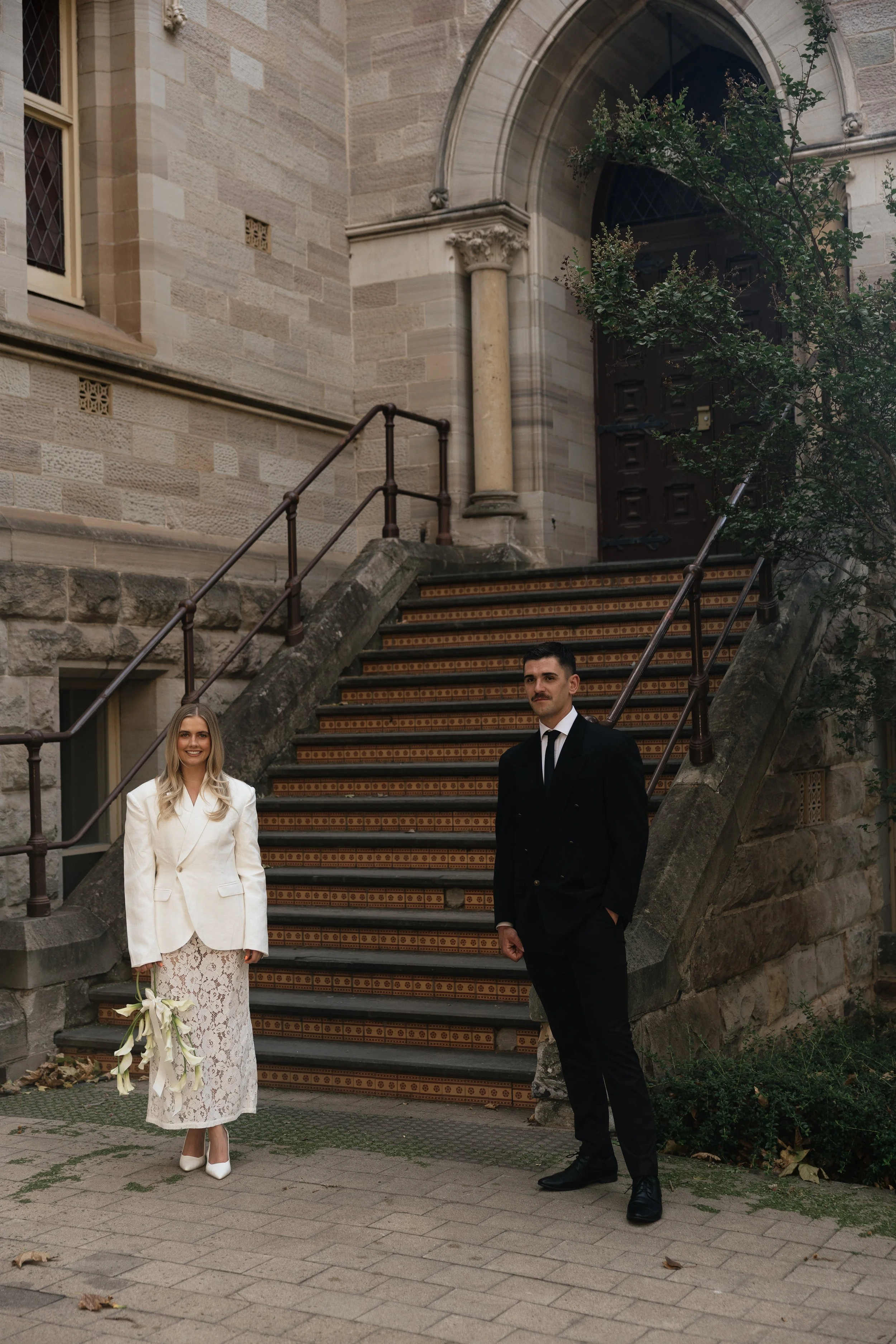 A woman in a white blazer and lace skirt holding a bouquet of white flowers, and a man in a black suit, stand in front of a stone staircase and an arched wooden door at a historic building.