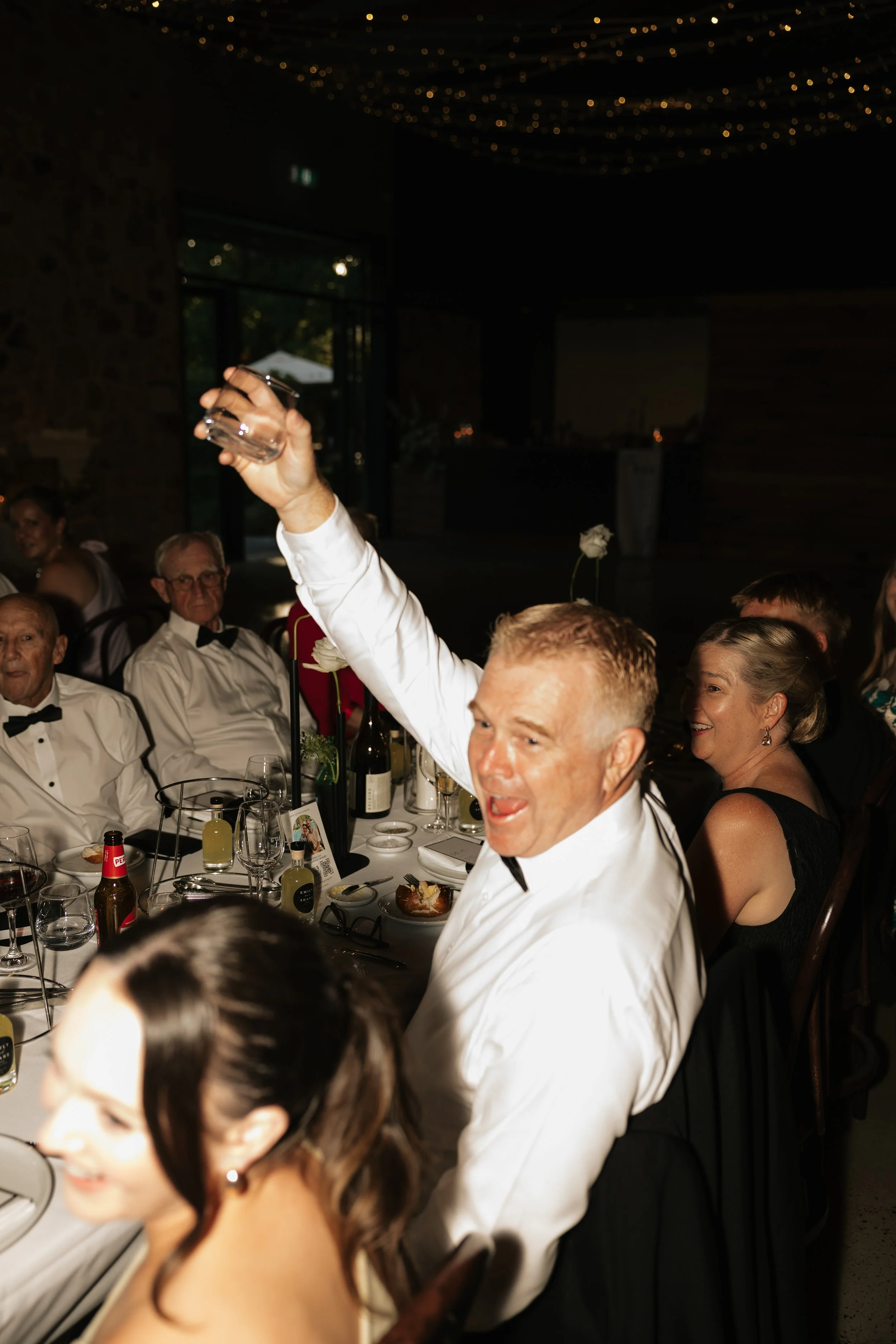 A man raising a glass at a formal dinner event with other guests seated at a round table, dressed in tuxedos and elegant attire, in a dimly lit room with string lights on the ceiling.