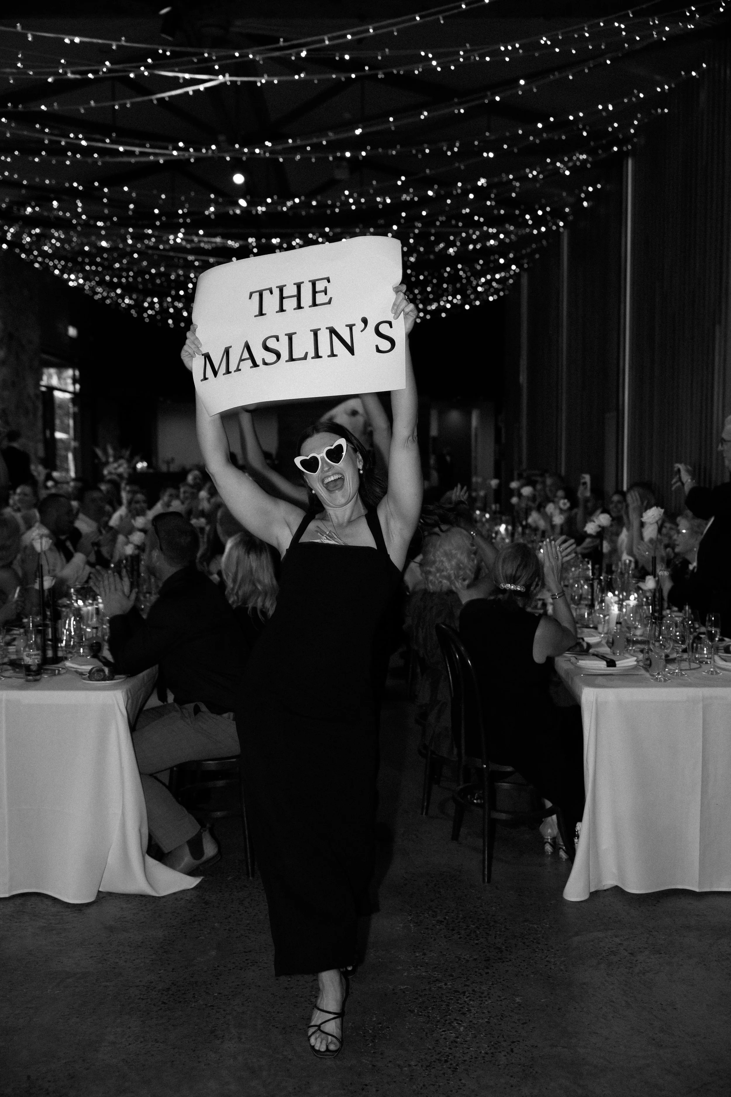 A woman wearing heart-shaped sunglasses and a black dress, holding a sign that reads 'THE MASLIN'S', smiling and dancing at a festive dinner event with decorated tables and string lights overhead.