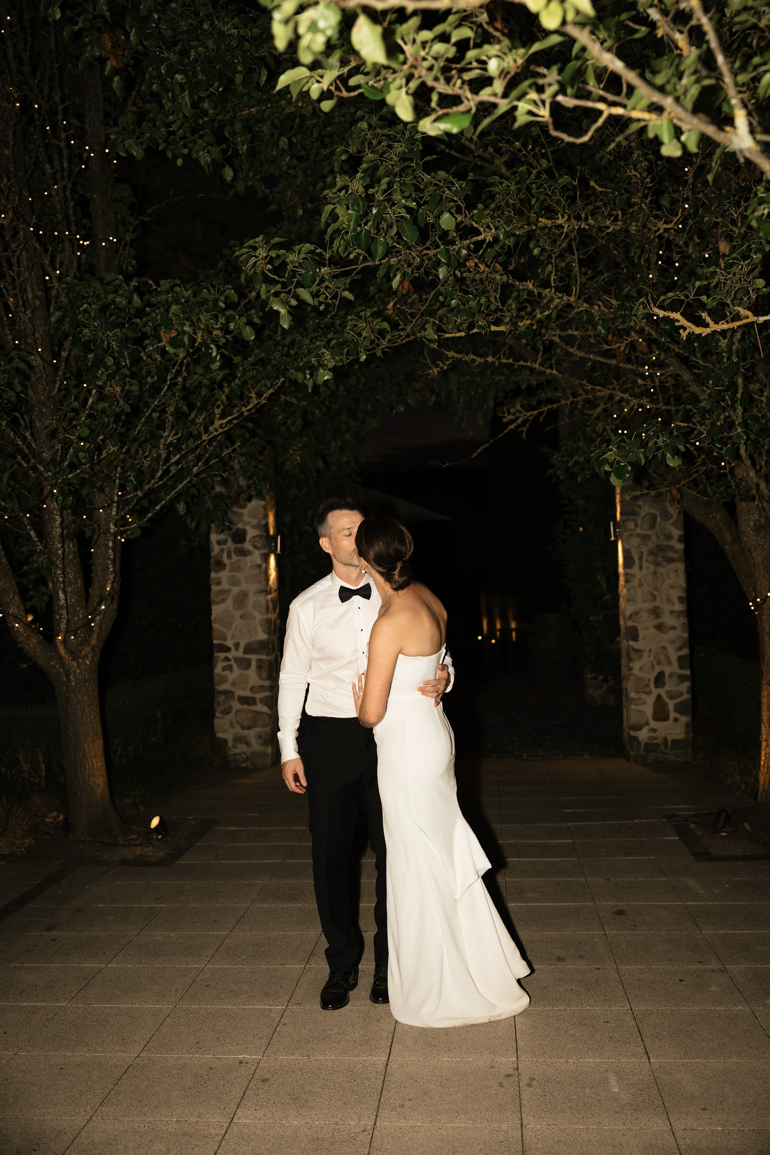 A newlywed couple shares a kiss under a canopy of lit trees at night, with stone archways in the background.
