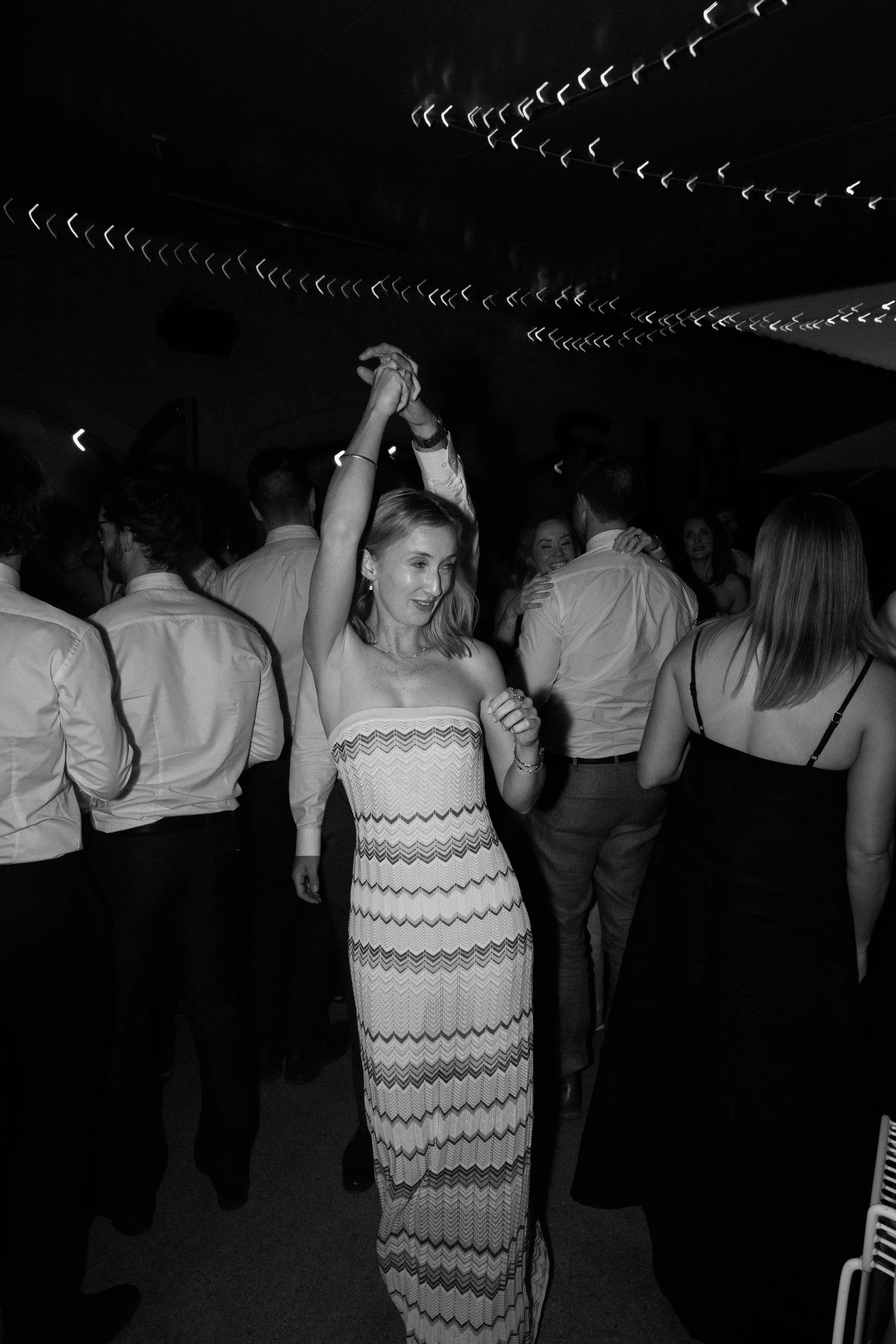 A woman dancing in a sleeveless, striped maxi dress at a party or dance event, surrounded by other people in formal or semi-formal attire. She has her arms raised and appears to be enjoying herself in a lively, dimly-lit setting with string lights ov