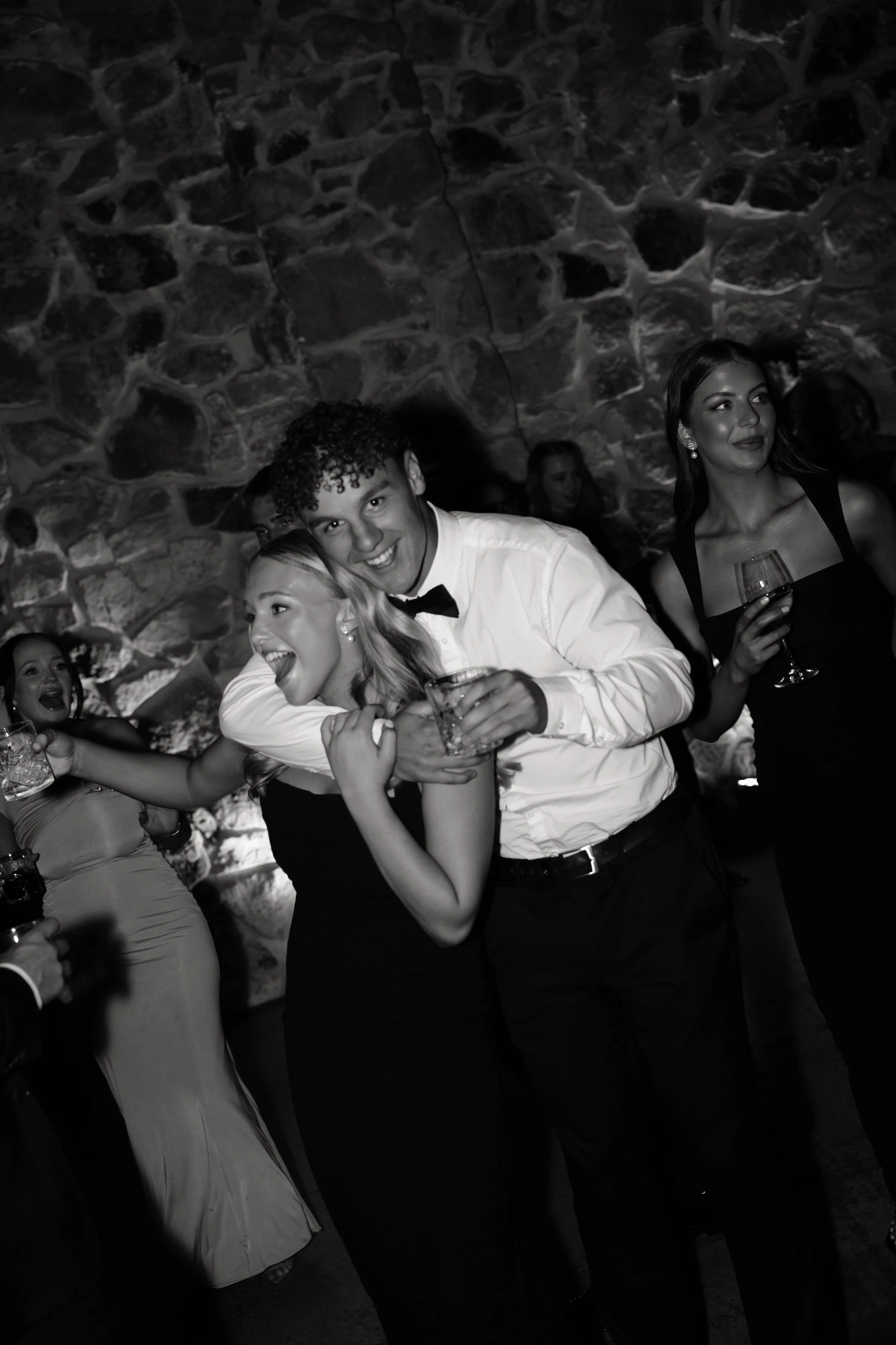 People dancing and celebrating at a party, with a stone wall in the background, in a black-and-white photo.