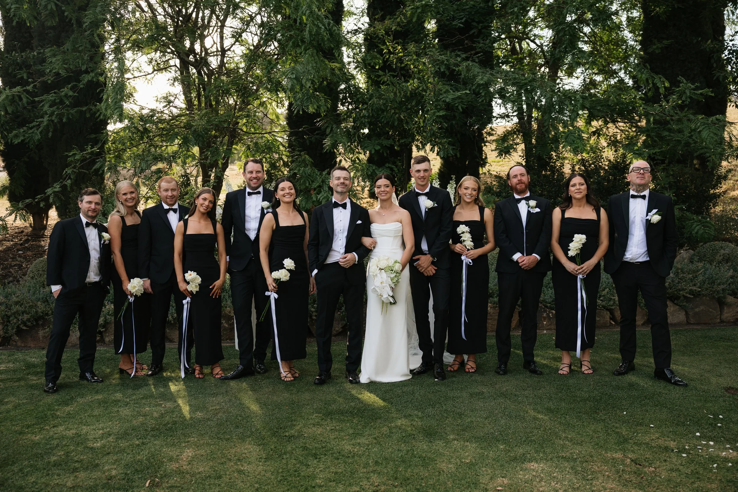 A group of 12 people, dressed in formal wedding attire, posing outdoors under trees. The group includes the bride and groom at the center, surrounded by friends or family. Golding Wines Wedding
