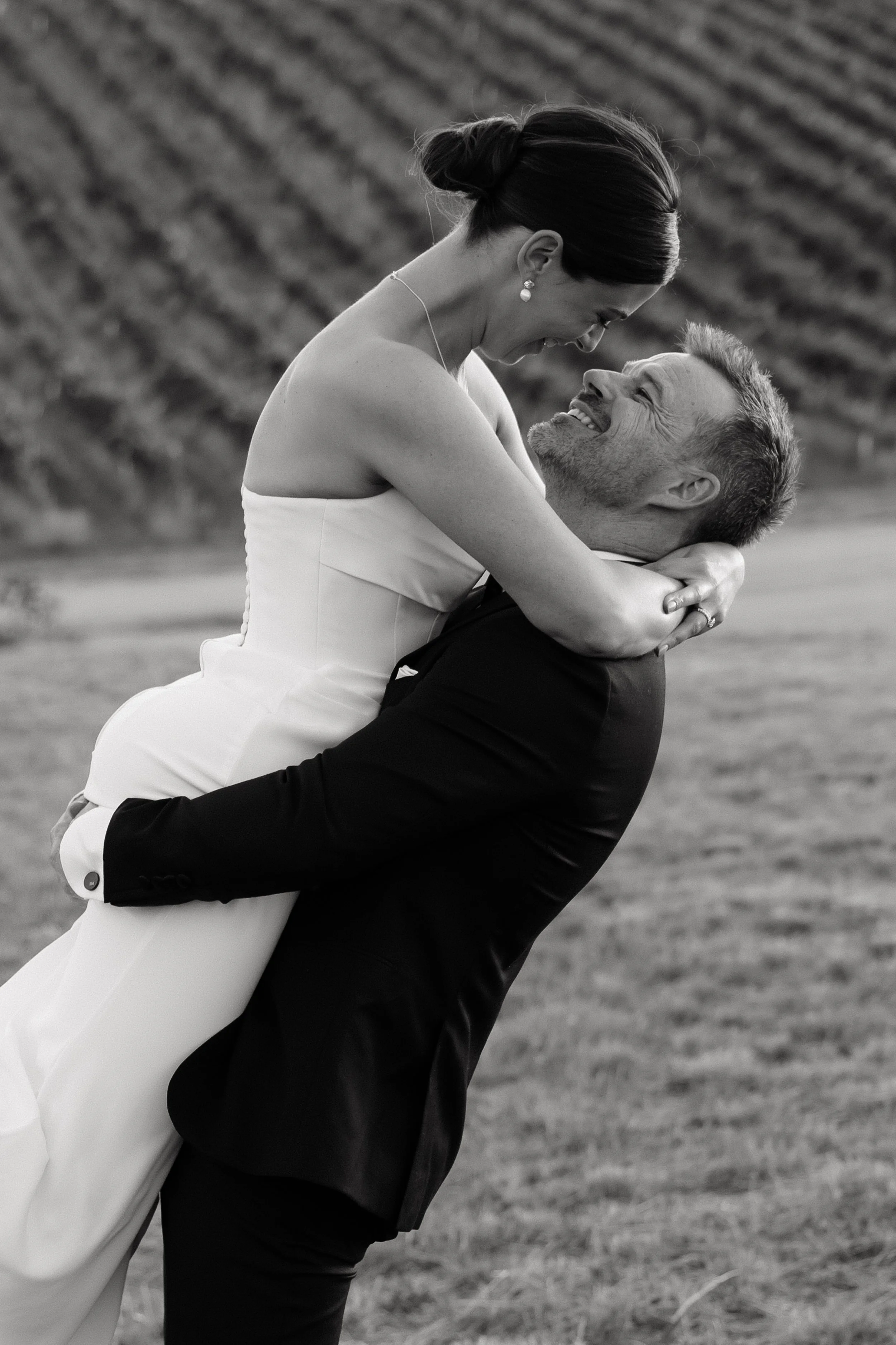 A black and white photo of a couple during their wedding, with the man lifting the woman who is wearing a wedding dress. They are smiling and looking at each other, with a blurred background of a field or garden. Golding Wines Wedding