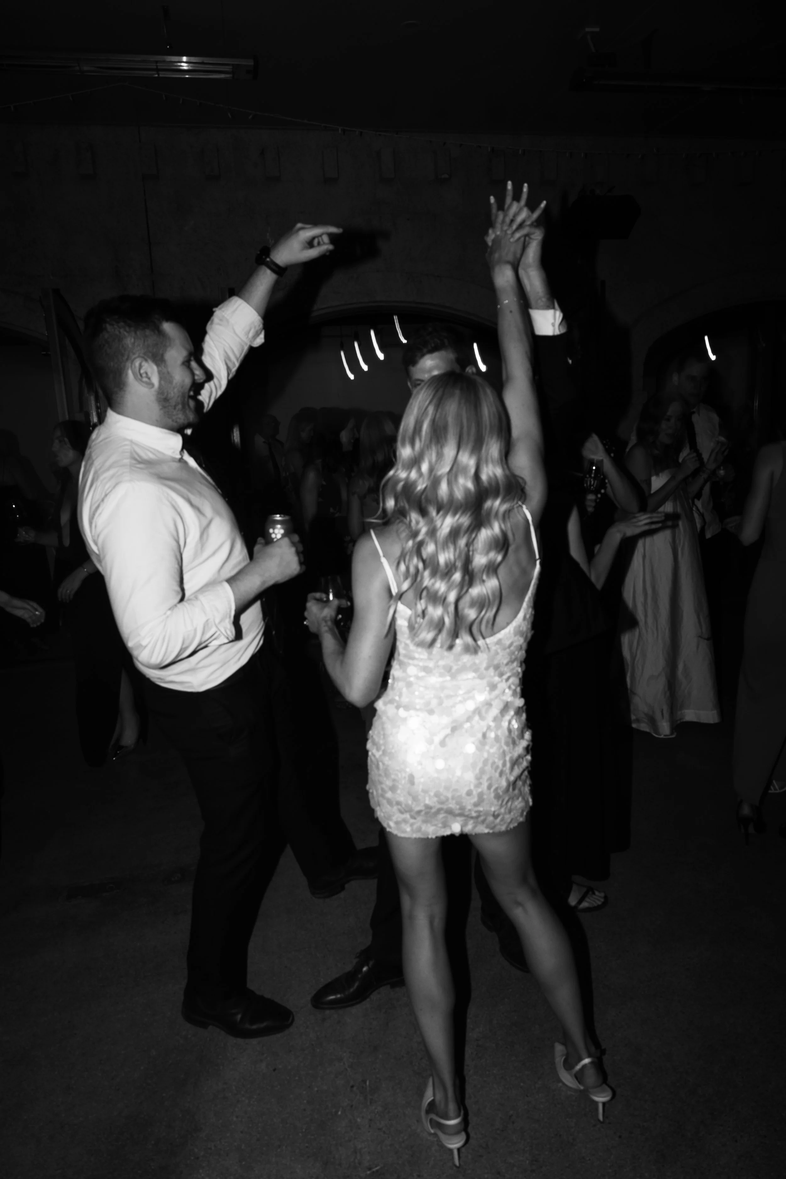 People dancing at a party, including a woman in a sparkly dress and a man in a shirt, in a dimly lit room.