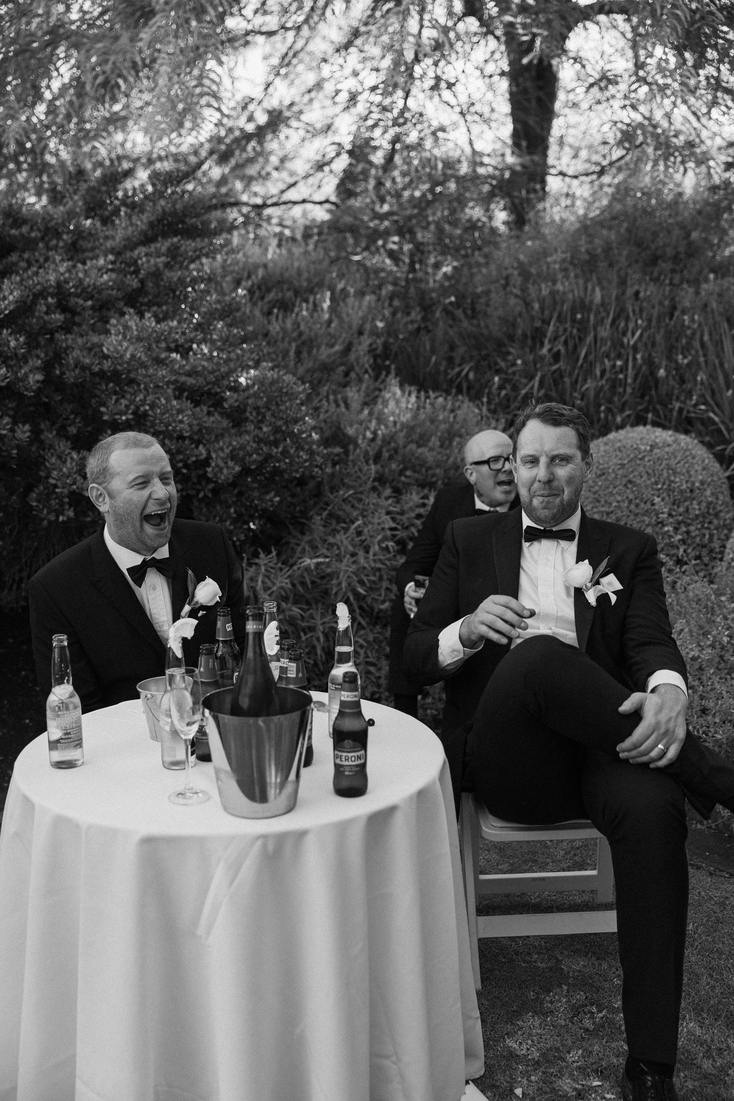 Three men in tuxedos sitting at a round table outdoors, two of them laughing and one smiling, with bottles of beer and glasses on the table, surrounded by trees and bushes.