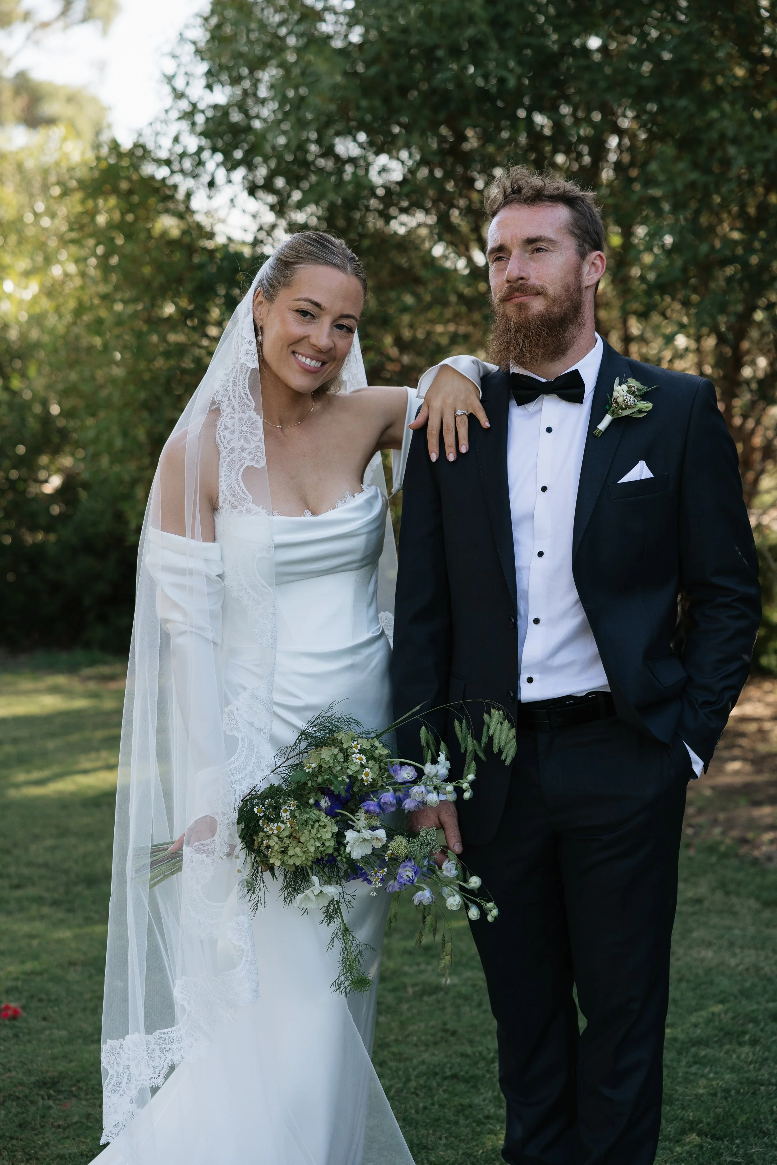 A bride and groom standing outdoors, smiling and posing for a photo during their wedding. The bride is wearing a white dress with lace details and a veil, holding a bouquet of flowers. The groom is dressed in a black tuxedo with a bow tie and a bouto